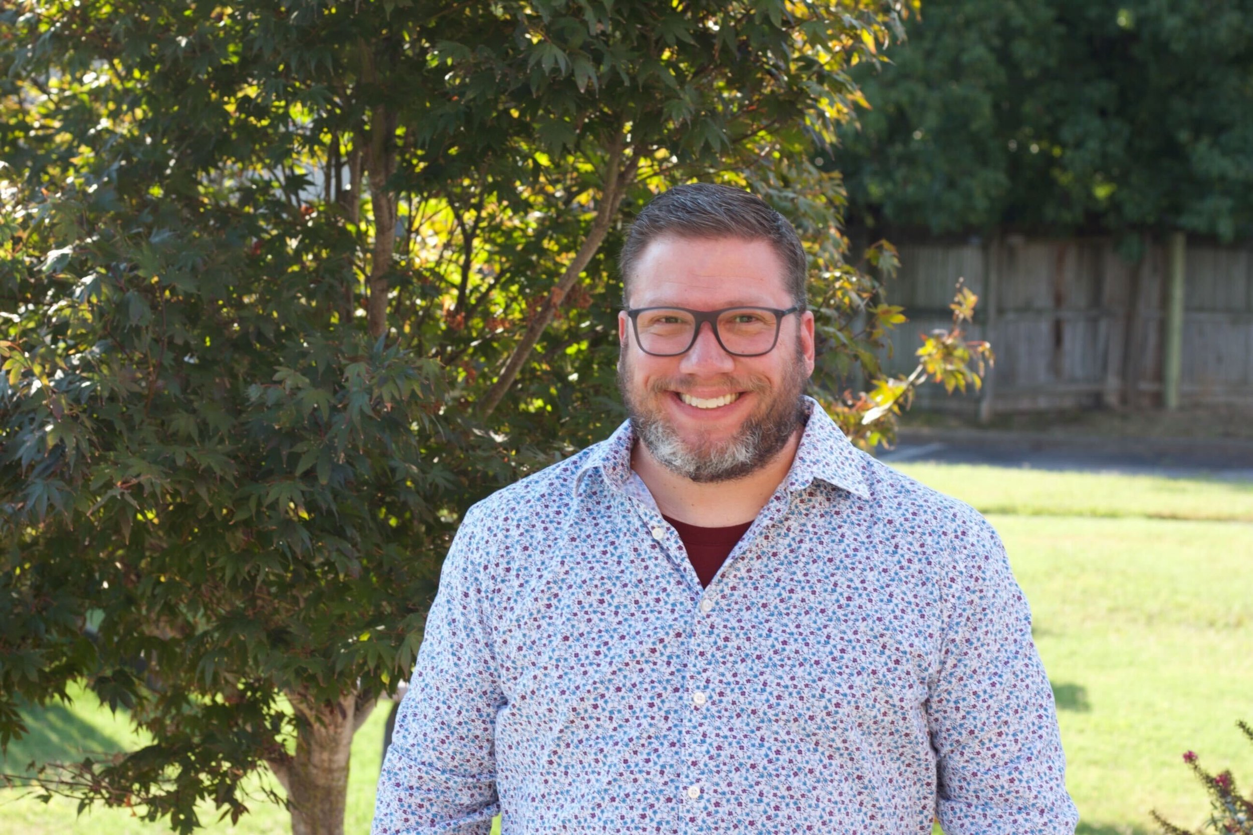 A smiling man with glasses and a beard wearing a floral shirt standing outdoors in front of trees and a wooden fence.