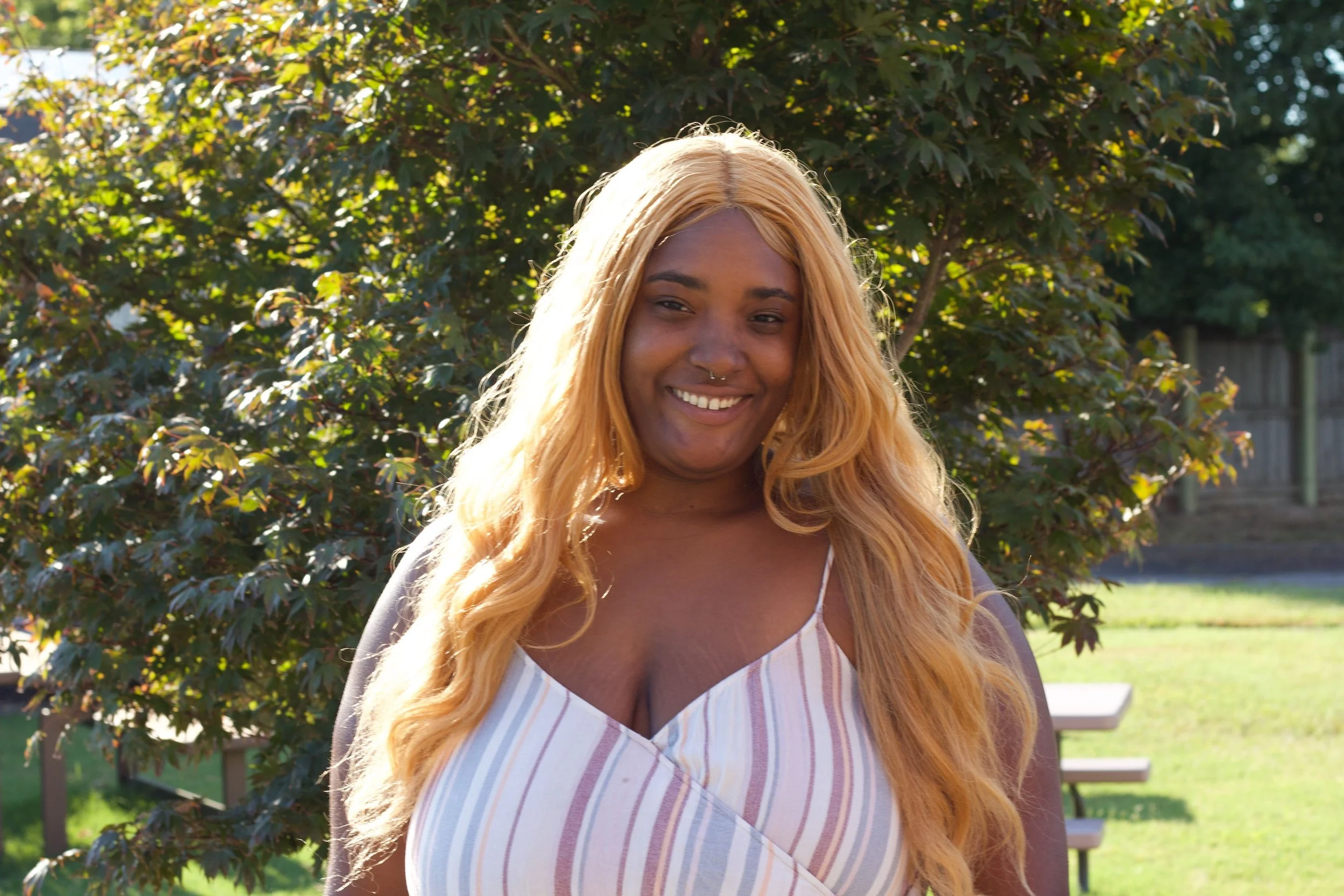 A woman with long, wavy blond hair smiling outdoors with a large bush and trees in the background.