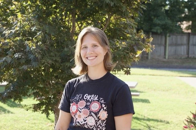 A young woman with shoulder-length hair smiling outdoors, standing in front of a leafy tree and a grassy yard.