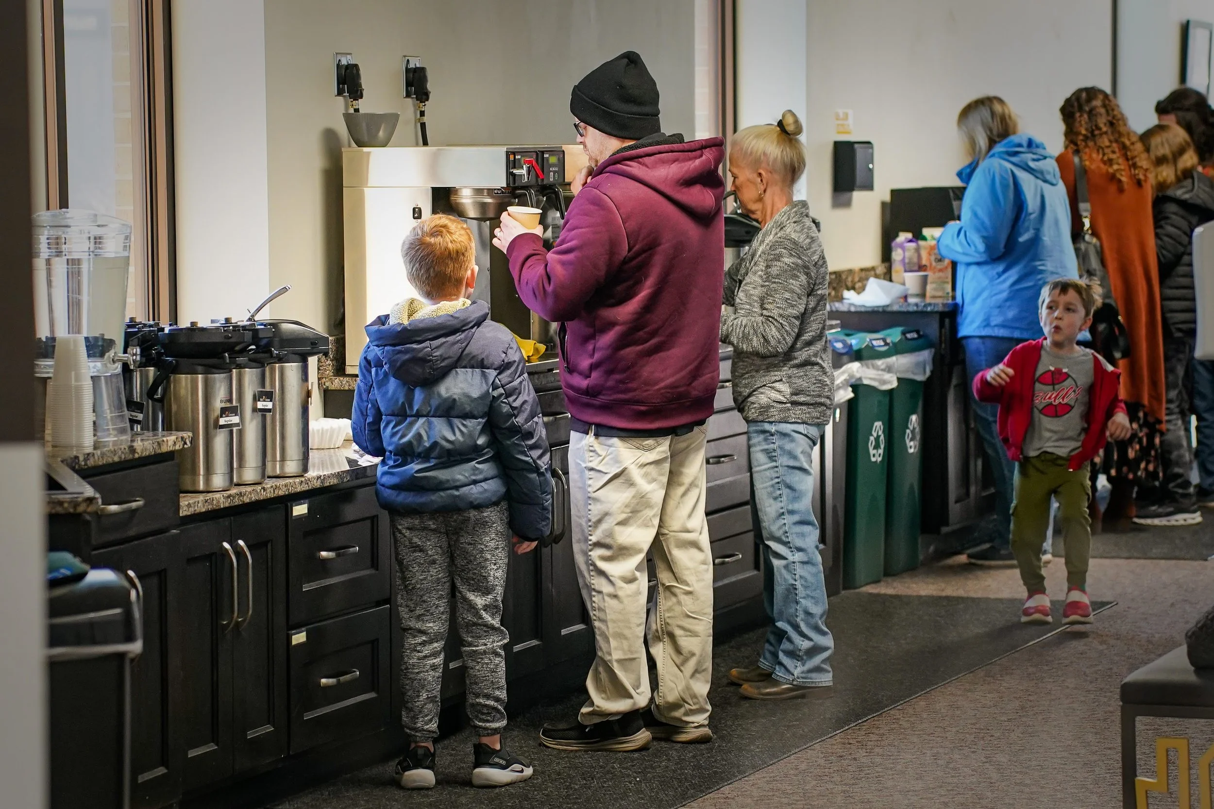 People standing in line at a coffee station in a cafe, with a man wearing a black beanie preparing a coffee for a boy.