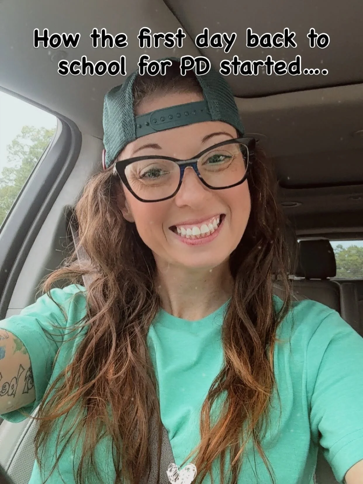 A woman with long, wavy brown hair, wearing glasses, a green cap backwards, and a turquoise shirt, smiles while sitting inside a vehicle, with text overlay that reads 'How the first day back to school for PD started...'.