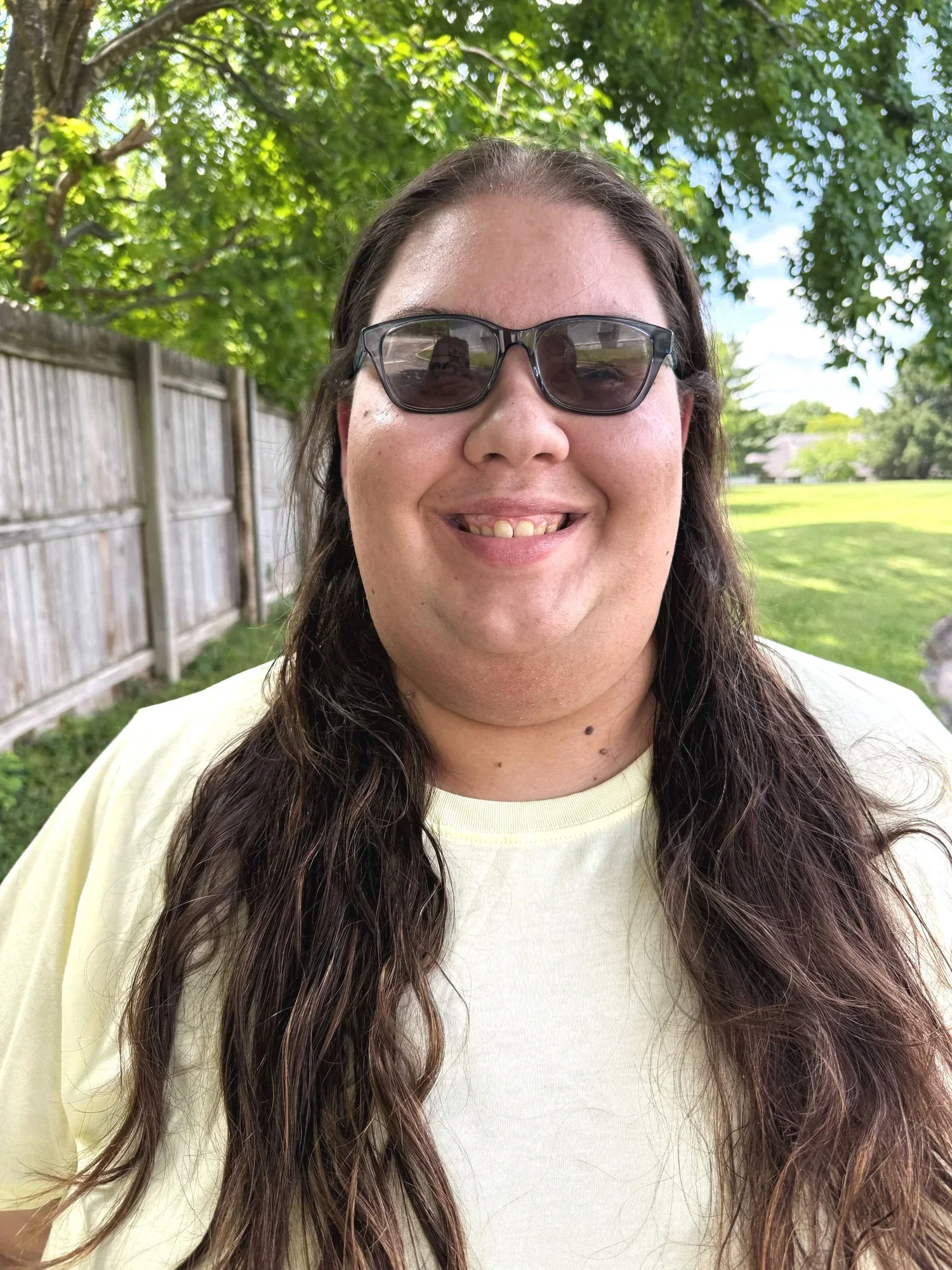 A person with long wavy brown hair wearing sunglasses and a light yellow shirt, smiling outdoors with a green leafy background and a wooden fence.
