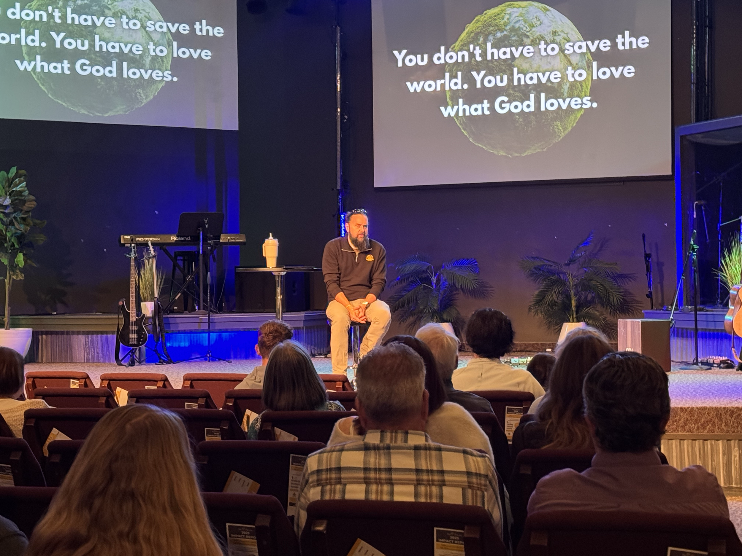 A man with a beard sitting on stage in front of large screens displaying a message about loving what God loves, with an audience seated in front of him.