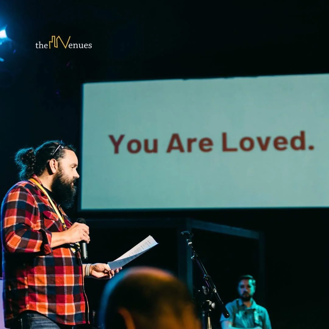 A man with a beard and long hair tied back, wearing a red plaid shirt, stands holding a microphone and reading from a paper, with a large screen behind him displaying the message "You Are Loved." in red letters. There are a few people seated in the audience.
