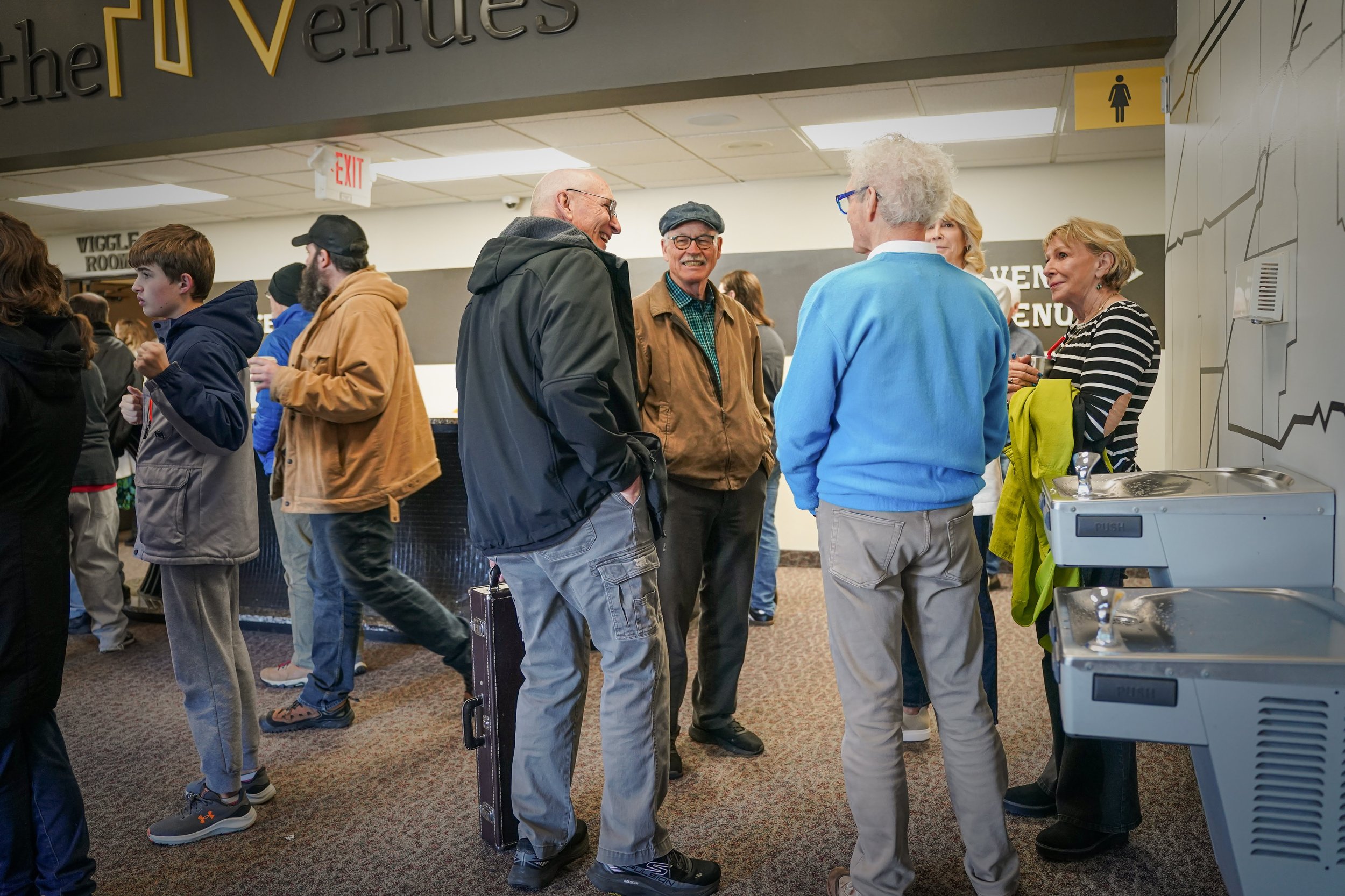 Group of people, mostly older adults, socializing and talking in line at an indoor setting, possibly a community center or event venue, with a water fountain station visible on the right.