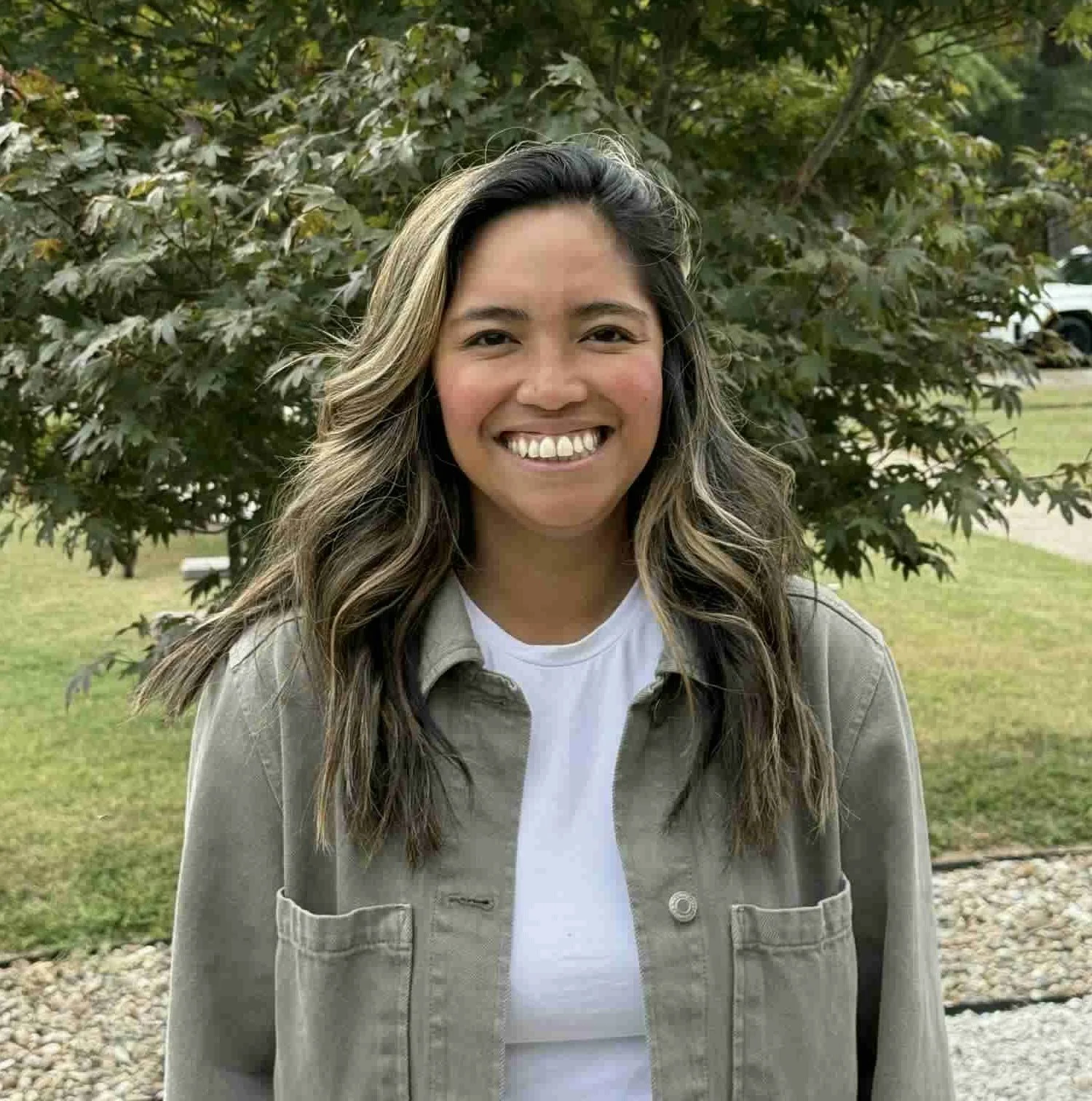 A woman with long wavy hair smiling outdoors in front of green foliage, wearing a white shirt and a beige jacket.