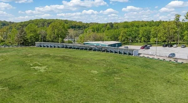 A long row of white covered picnic tables on a grassy field with a parking lot and cars in the background, surrounded by trees under a partly cloudy sky.