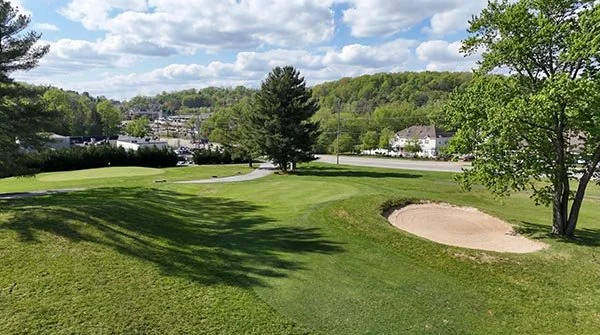 A golf course with lush green grass, sand bunker, trees, and a view of houses and hills under a partly cloudy sky.