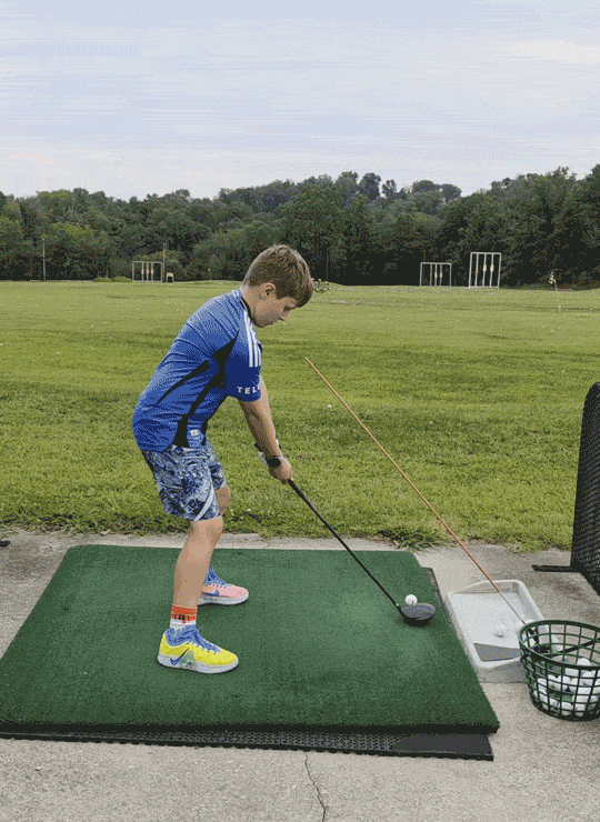 A young boy practicing golf at an outdoor driving range, wearing a blue sports shirt, patterned shorts, and colorful shoes, holding a golf club ready to hit a golf ball.