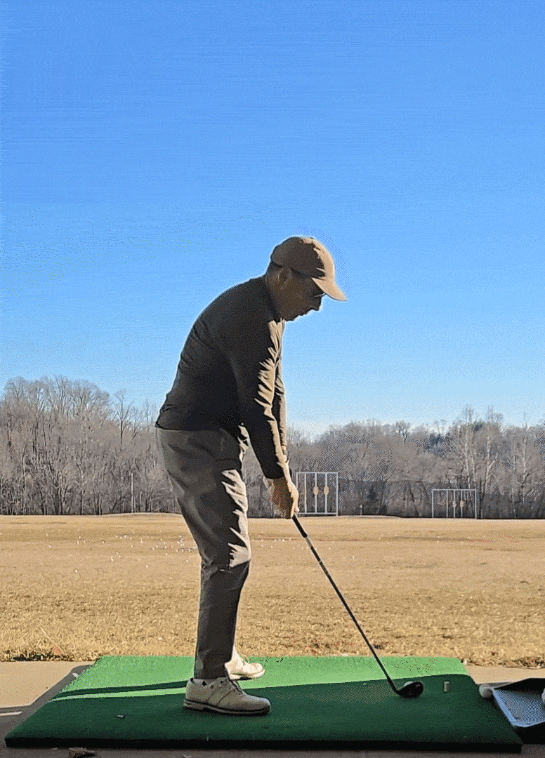 Person practicing golf swing on a driving range with golf clubs, wearing a cap, dark jacket, gray pants, sneakers, and a green mat, under a clear blue sky.