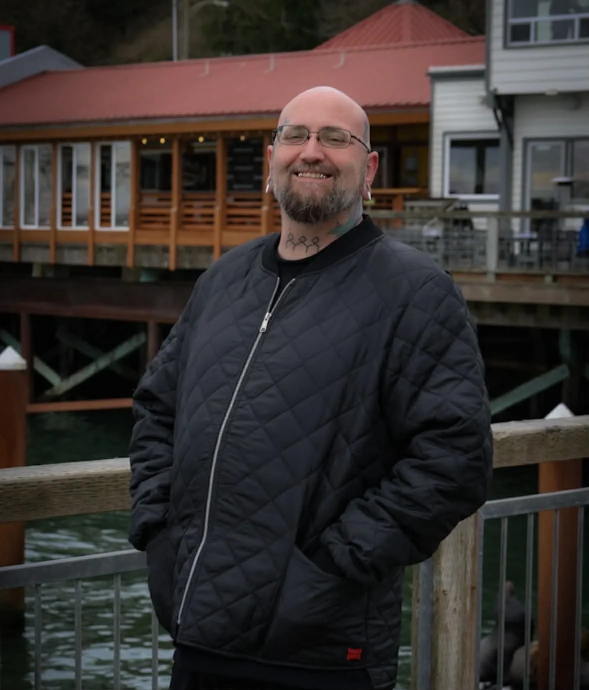 A smiling man with glasses, a beard, and tattoos on his neck, wearing a black quilted jacket, standing on a wooden dock near water with houses and a red-roofed building in the background.
