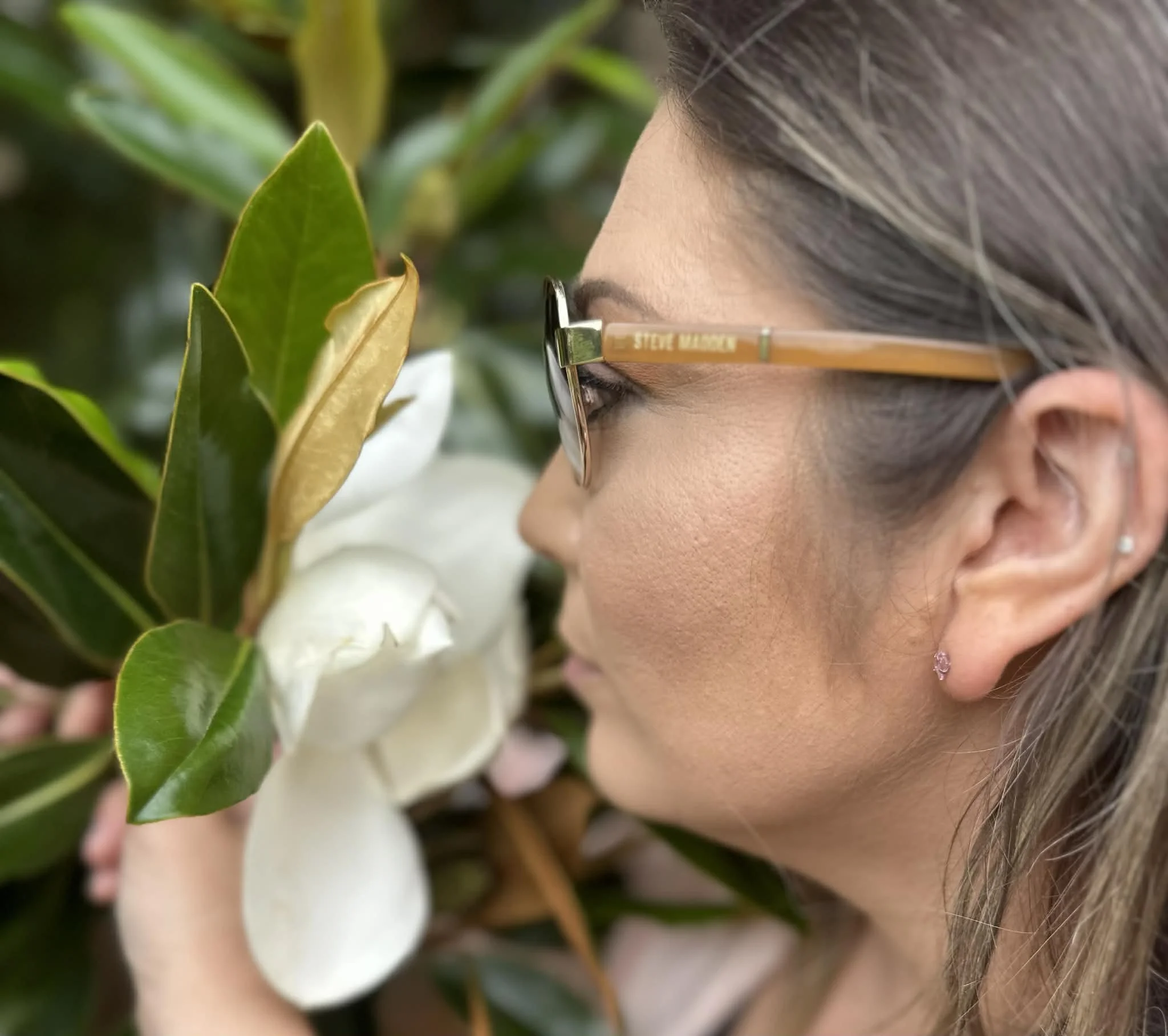 A woman with glasses and earrings smelling a white flower surrounded by green leaves.