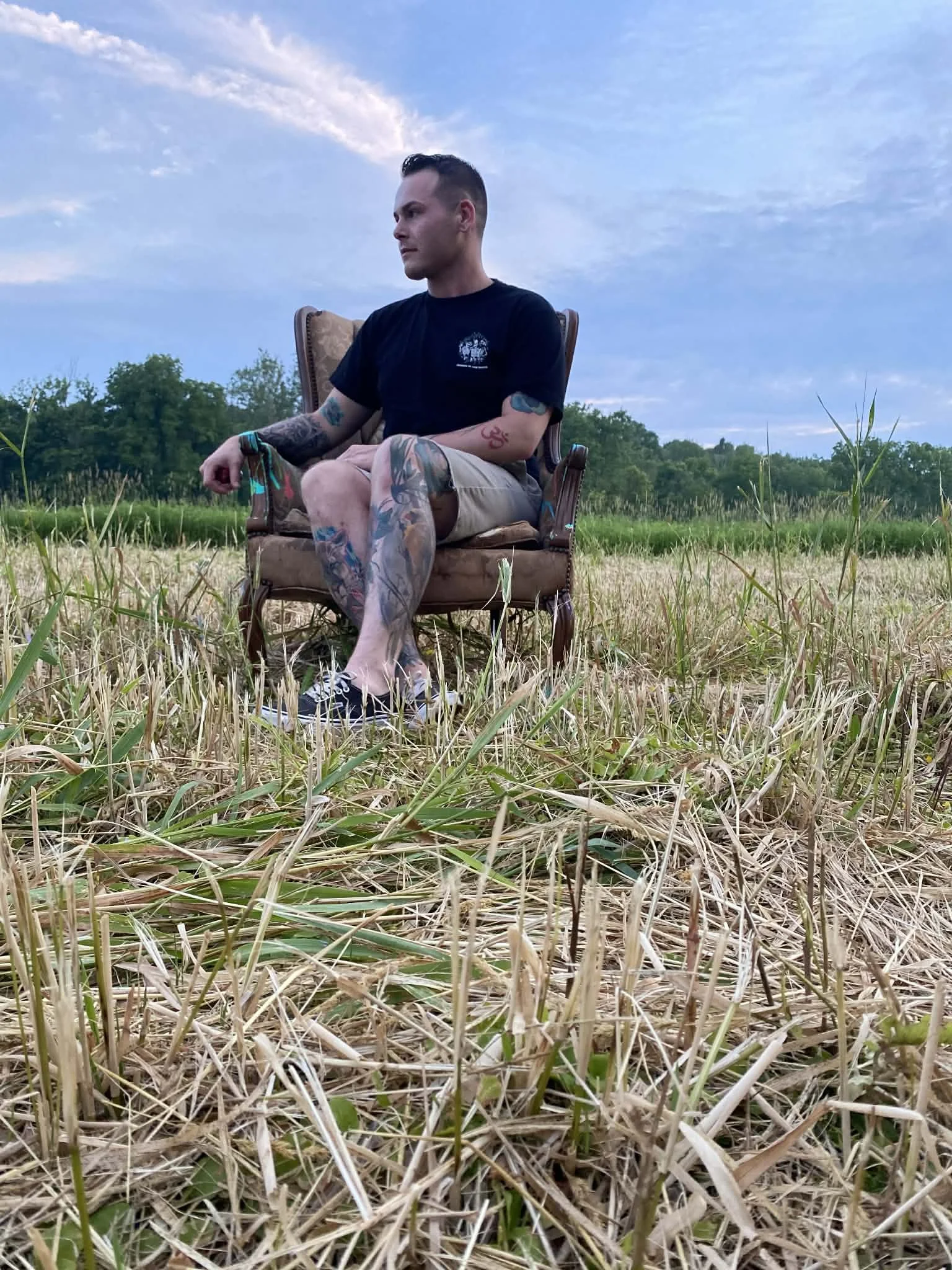 A man with tattoos sitting cross-legged on a vintage armchair in a field of dry grass, with trees and a cloudy sky in the background.