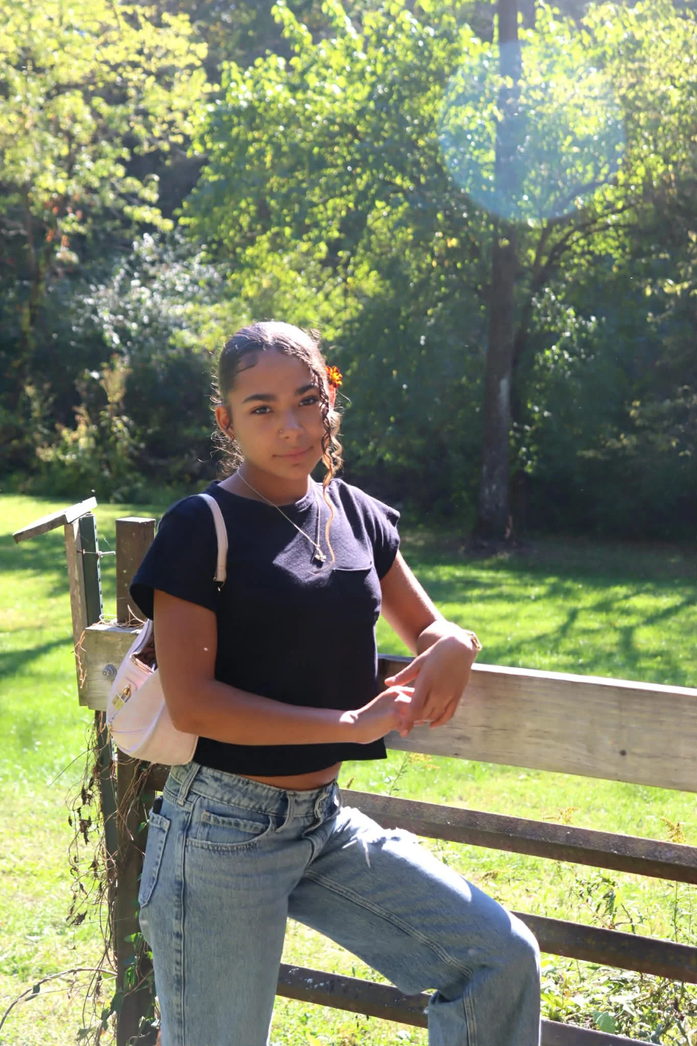 Young girl standing outdoors near a wooden fence, with green trees and sunlight in the background.