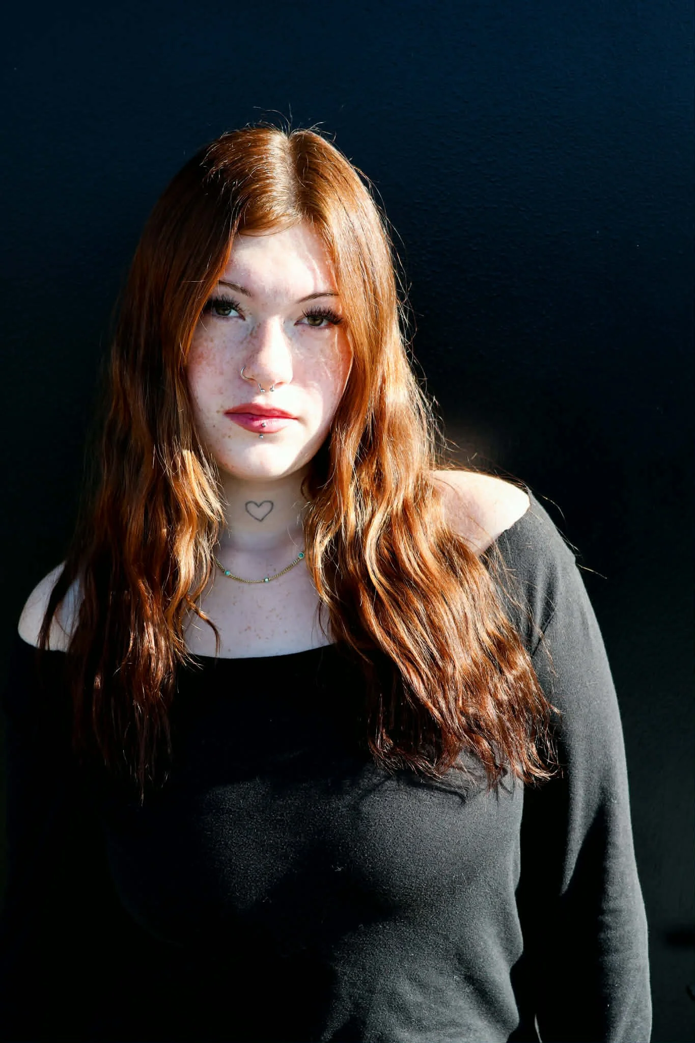 A young woman with long wavy red hair, pale skin with freckles, wearing a black off-shoulder top, a small necklace, and face piercings, standing against a dark background.