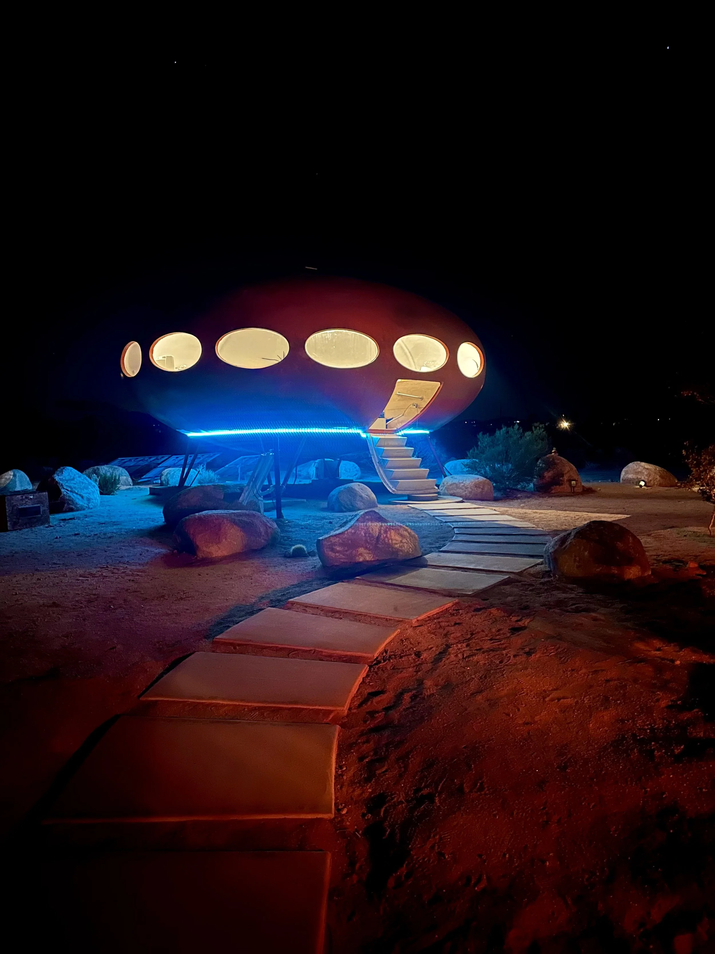 A futuristic house with an oval shape, circular windows, illuminated by colorful lights at night, surrounded by rocks and a pathway with stepping stones.