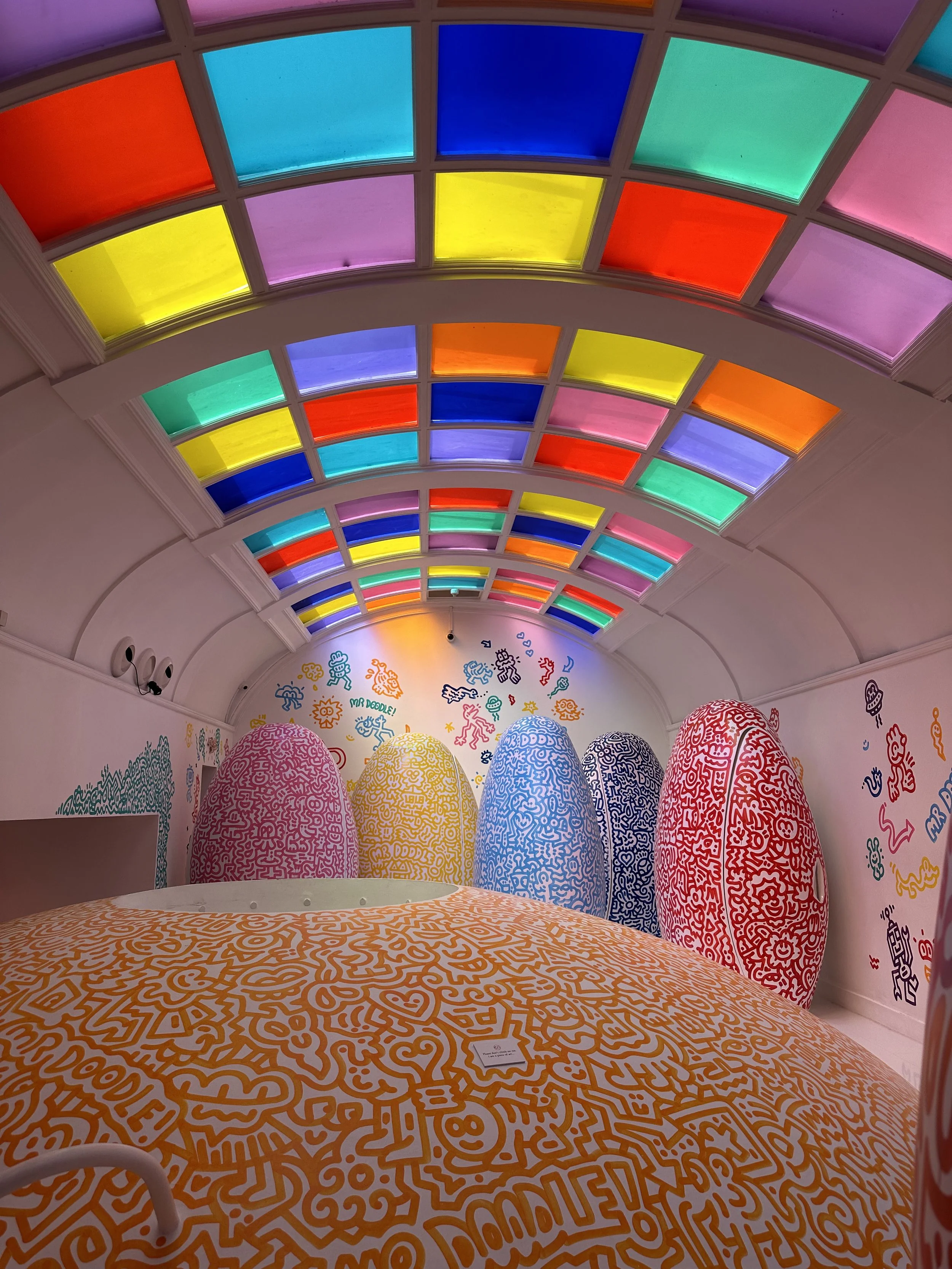 Colorful stained glass ceiling with a rainbow pattern, prominent orange, yellow, blue, green, purple, and pink panes, in an art installation space with large decorative worm-shaped sculptures covered in doodles and graffiti-style artwork on the walls.