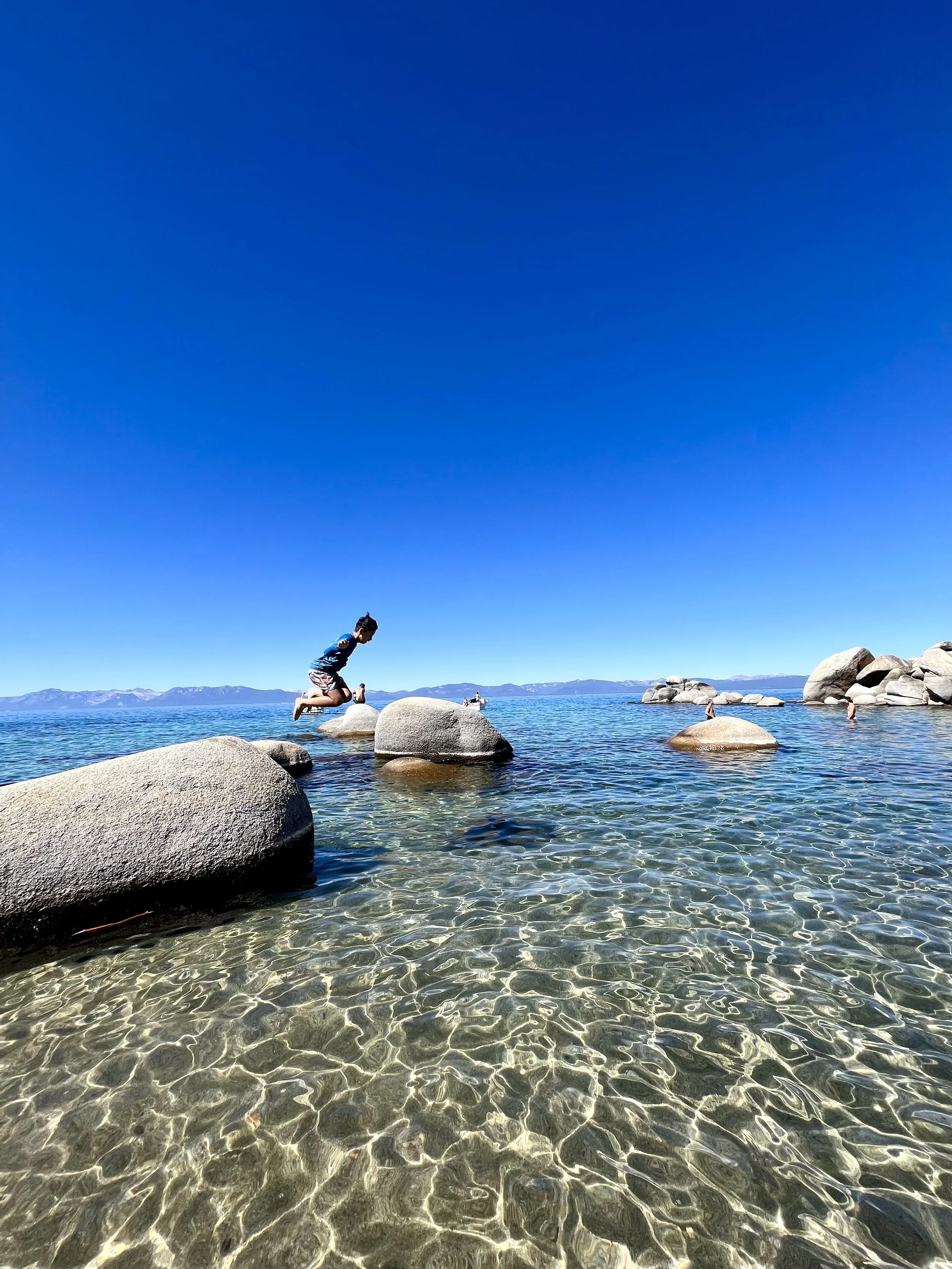 Child jumping from a large rock into clear water at a lakeside, with rocks and mountains in the background under a blue sky.