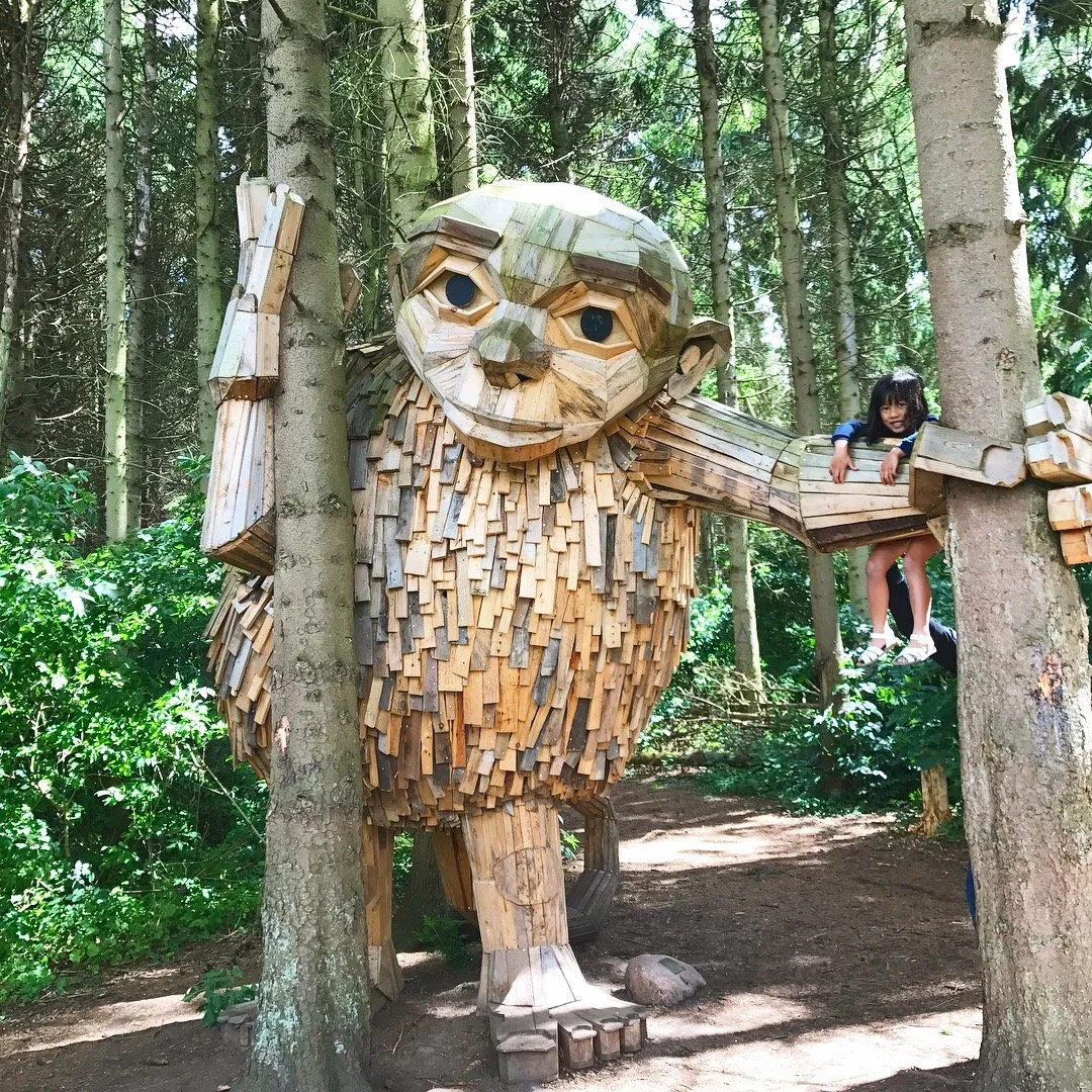 A large wooden sculpture of a monkey is placed in a forest, with a child sitting on its outstretched arm.