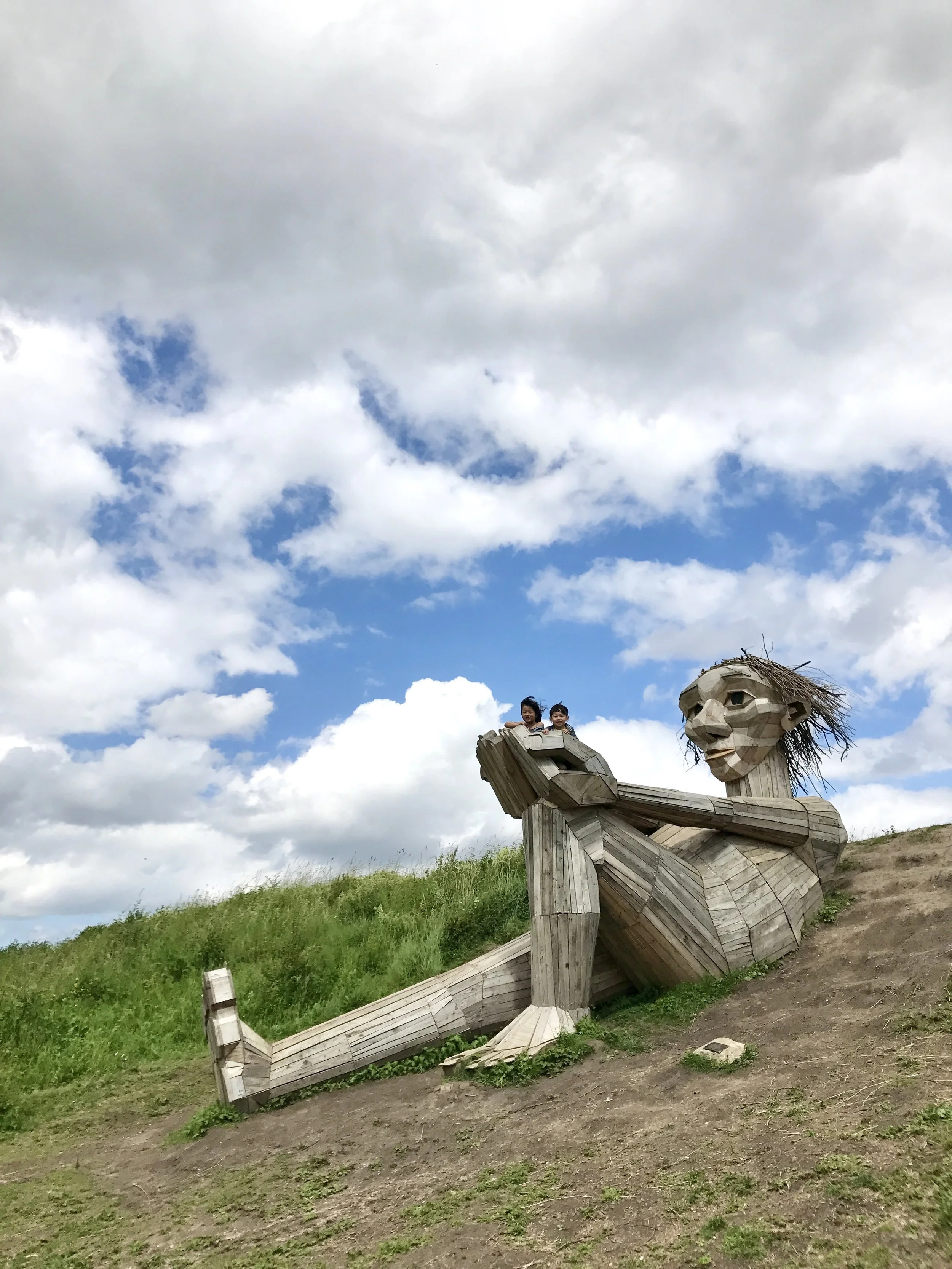 Wooden sculpture of a dragon with children sitting on its head in an outdoor area with grass and dirt. The sky is partly cloudy with blue patches.