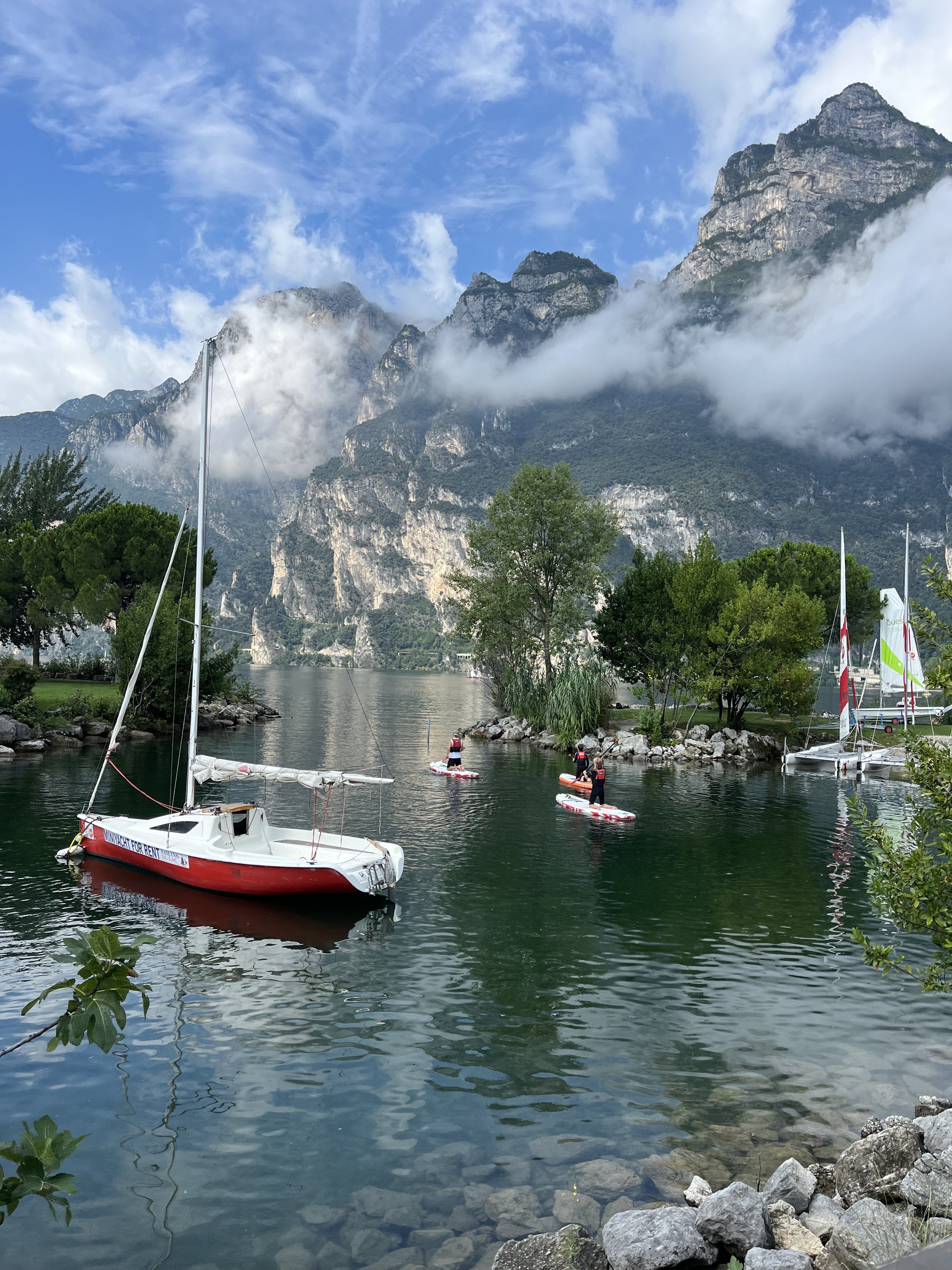 A lakeside scene with sailboats and paddleboarders, surrounded by green trees and rocky shores, with tall mountain cliffs partly covered in mist and clouds under a blue sky with wispy clouds.