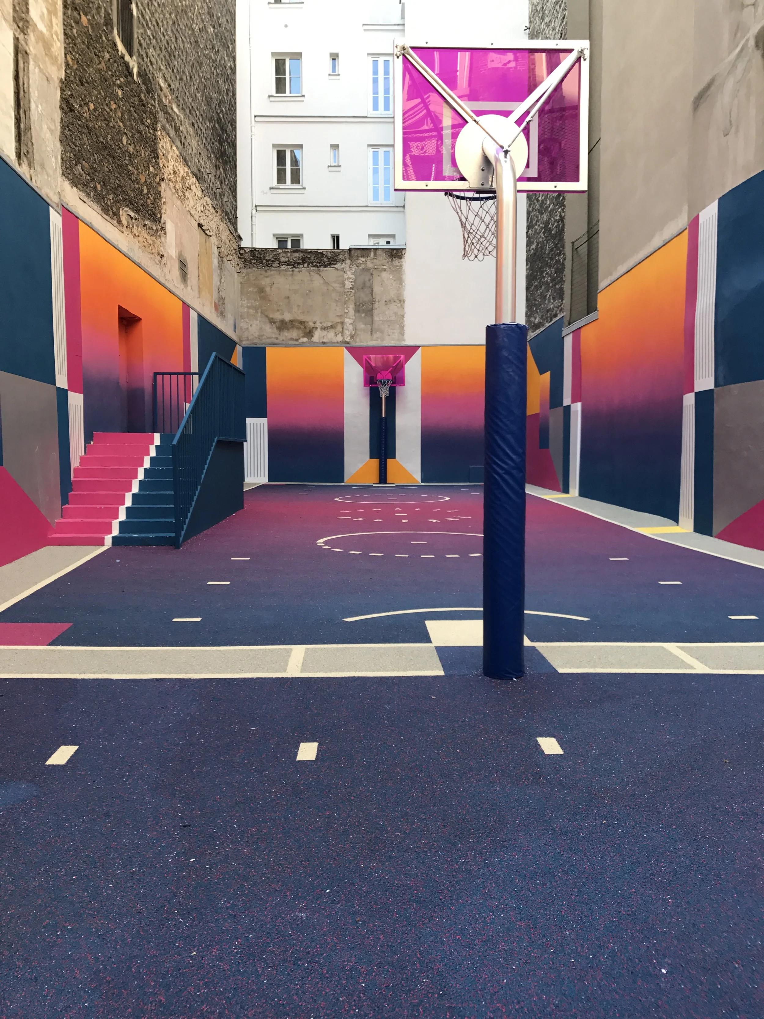 An urban basketball court with vibrant, colorful wall art and two basketball hoops, one of which has a purple backboard, surrounded by tall buildings.