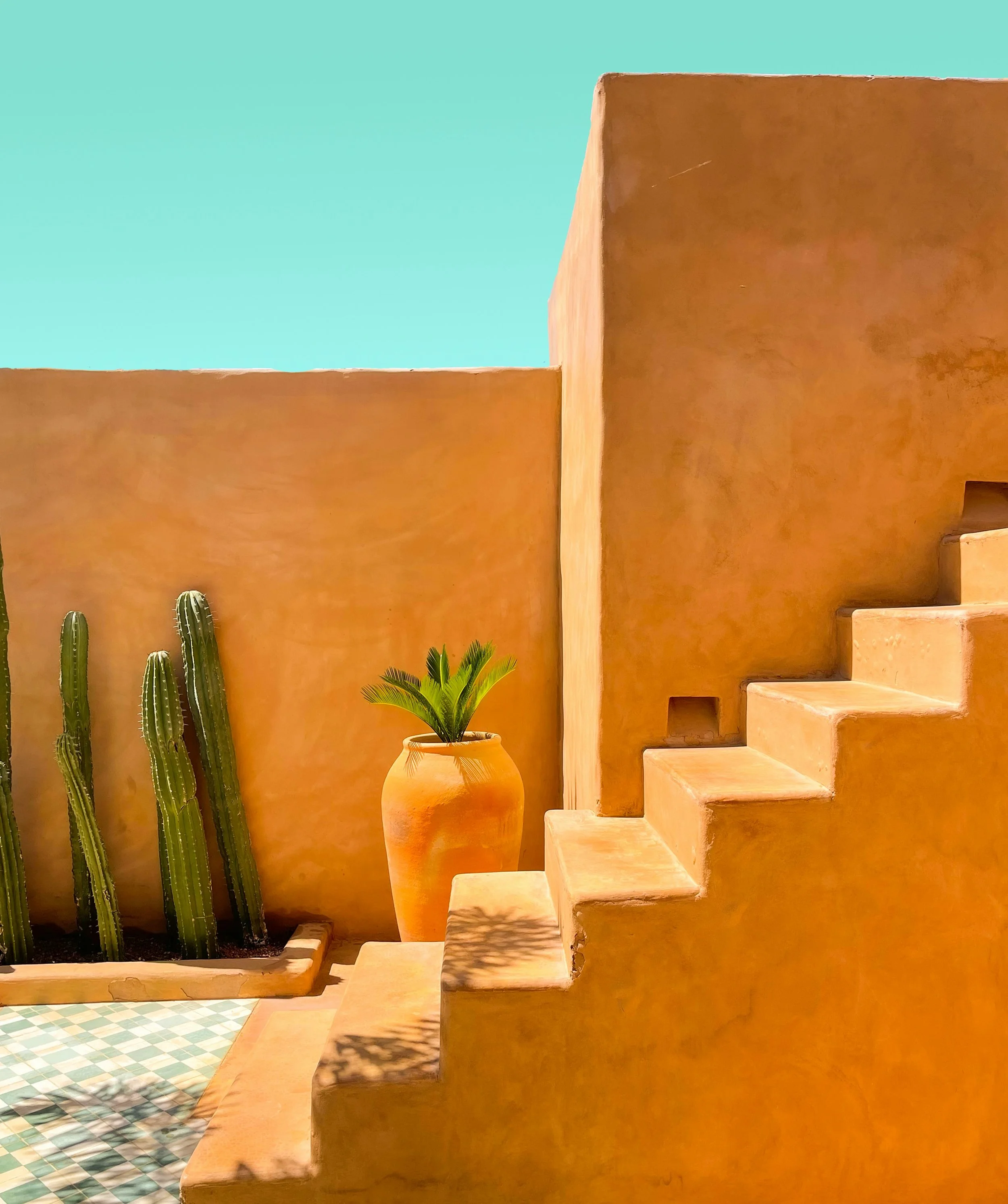 Terracotta-colored staircase outside with a tall terracotta pot holding a green plant, and a tiled courtyard with green cactuses, against a turquoise sky.