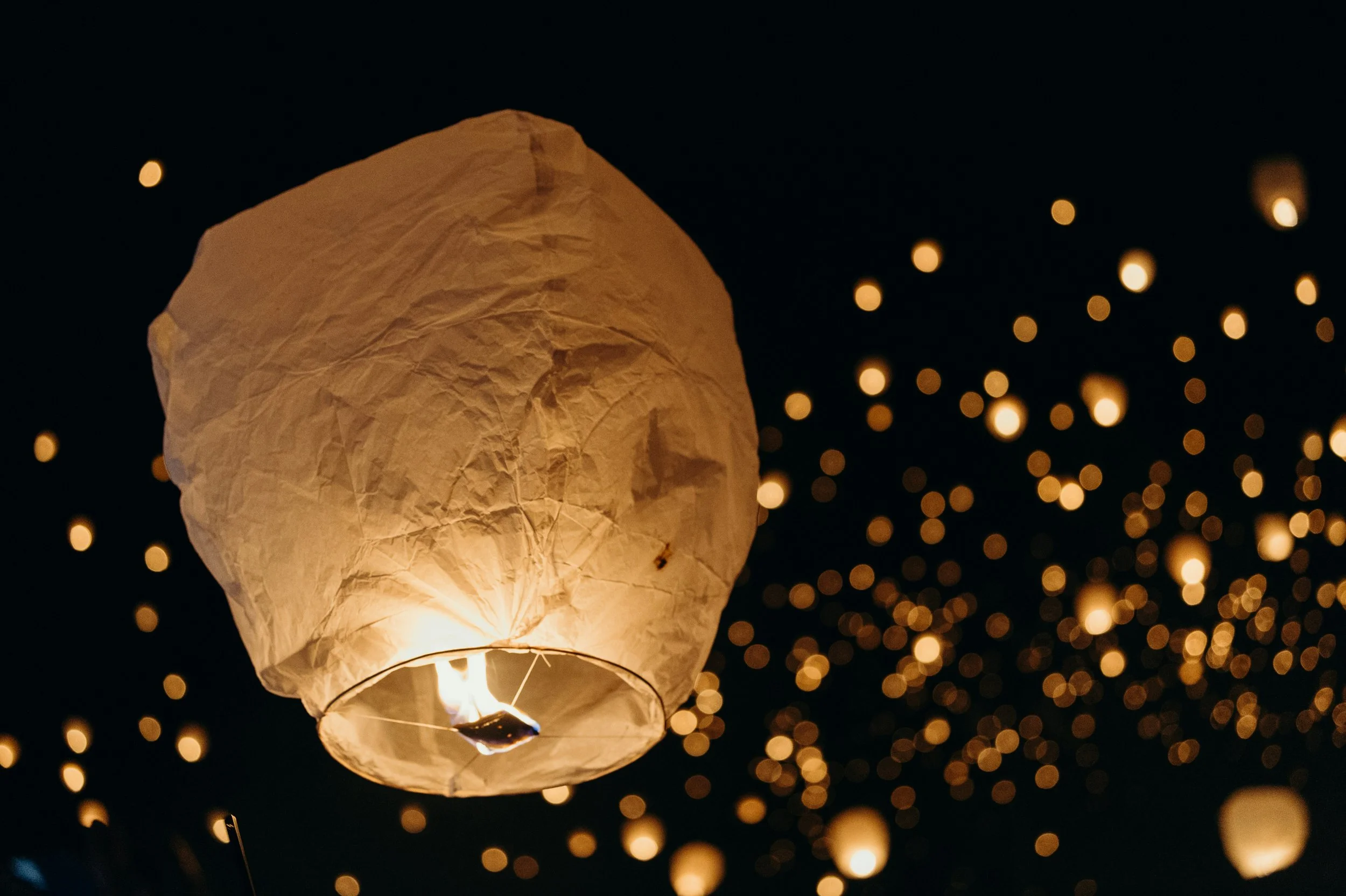 A paper lantern glowing with light against a dark night sky, surrounded by small blurred golden lights.