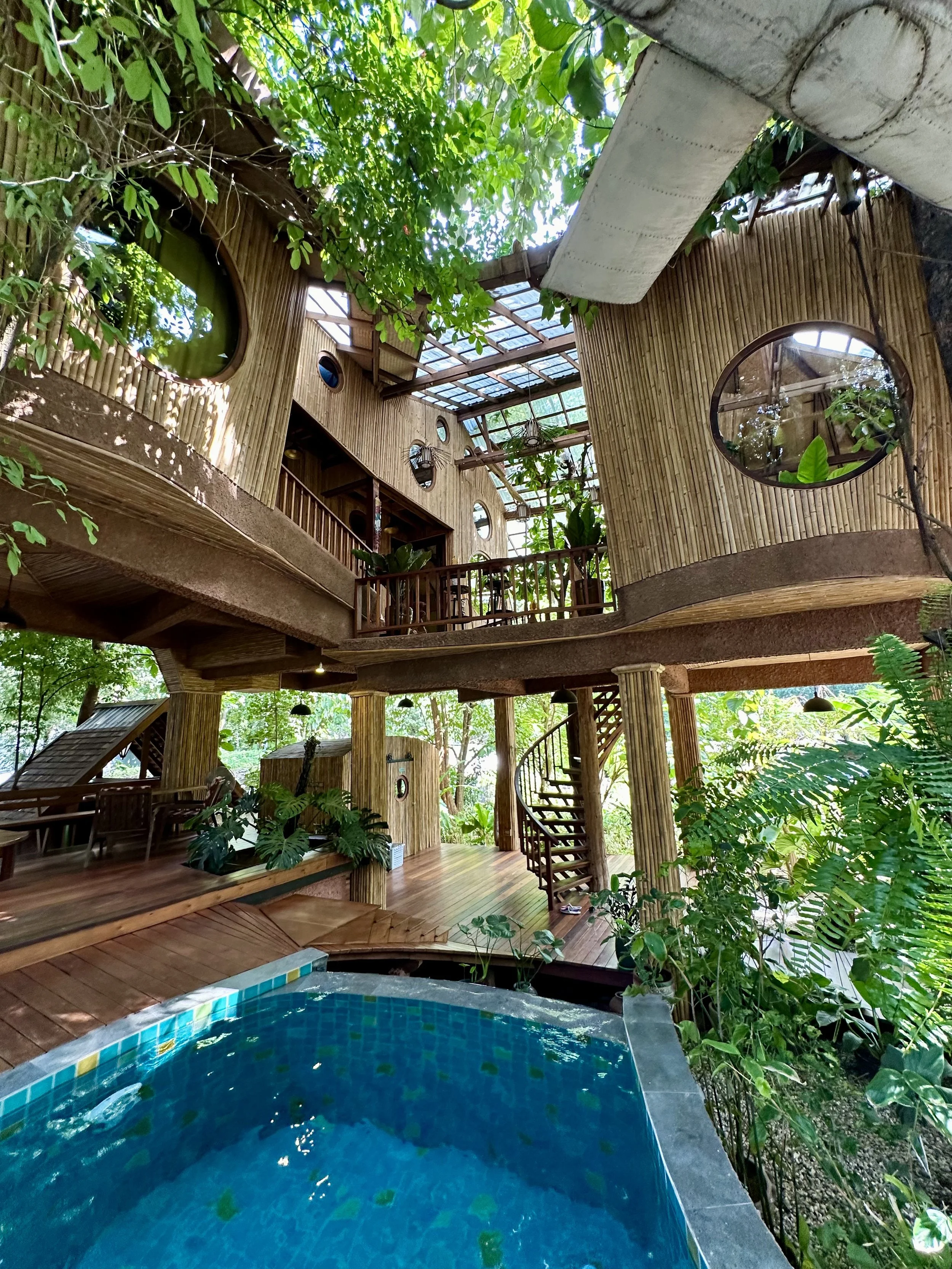 A multi-story treehouse built with bamboo and wood, surrounded by lush green foliage. The treehouse features round windows, a balcony with wooden railings, and a staircase. A partial view of a swimming pool with blue tiles is visible in the foreground.