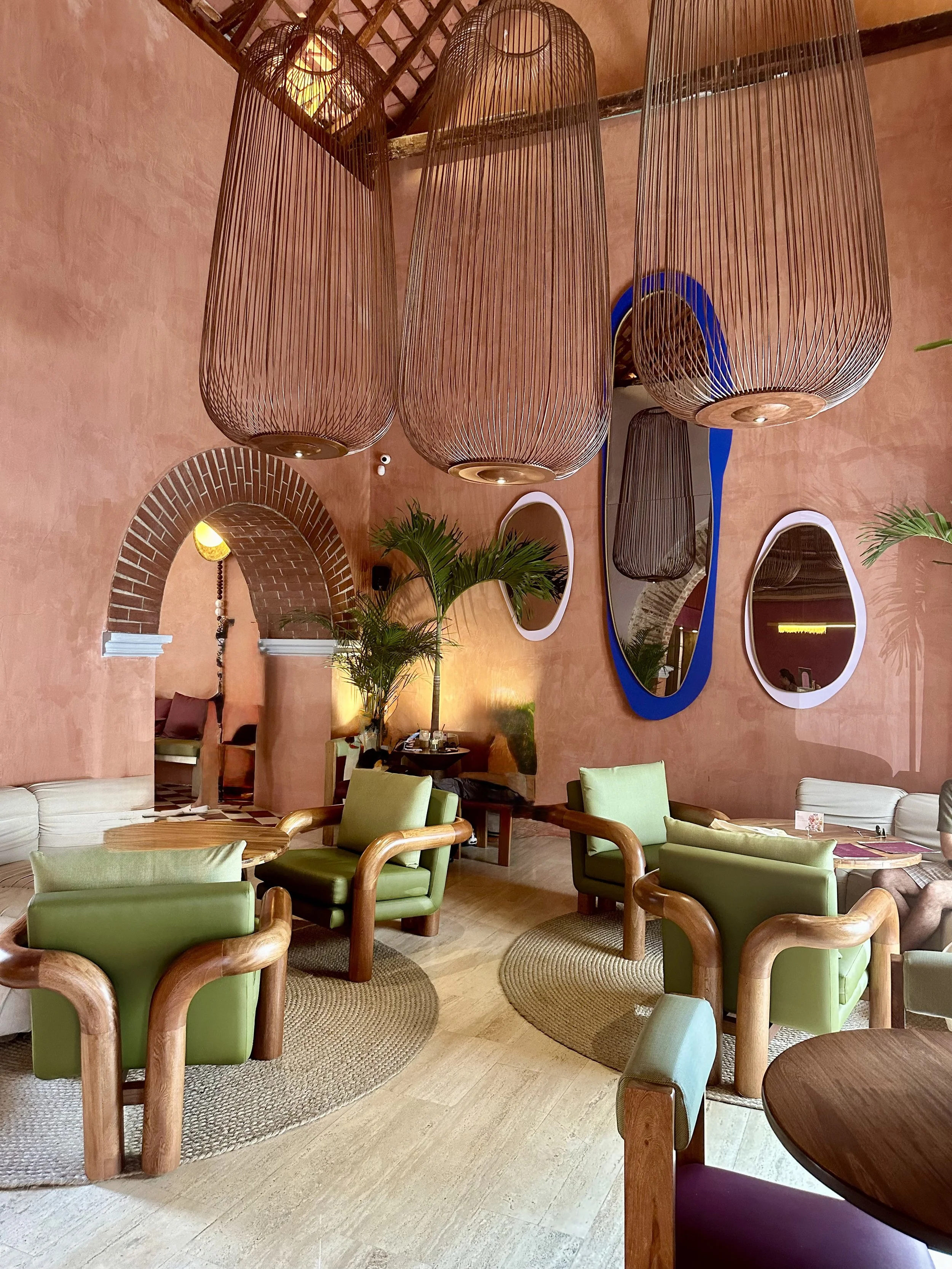 A cozy indoor lounge area with green armchairs, wooden tables, and a textured beige rug. Multiple woven pendant lights hang from the ceiling, and there are decorative mirrors and plants on the walls.