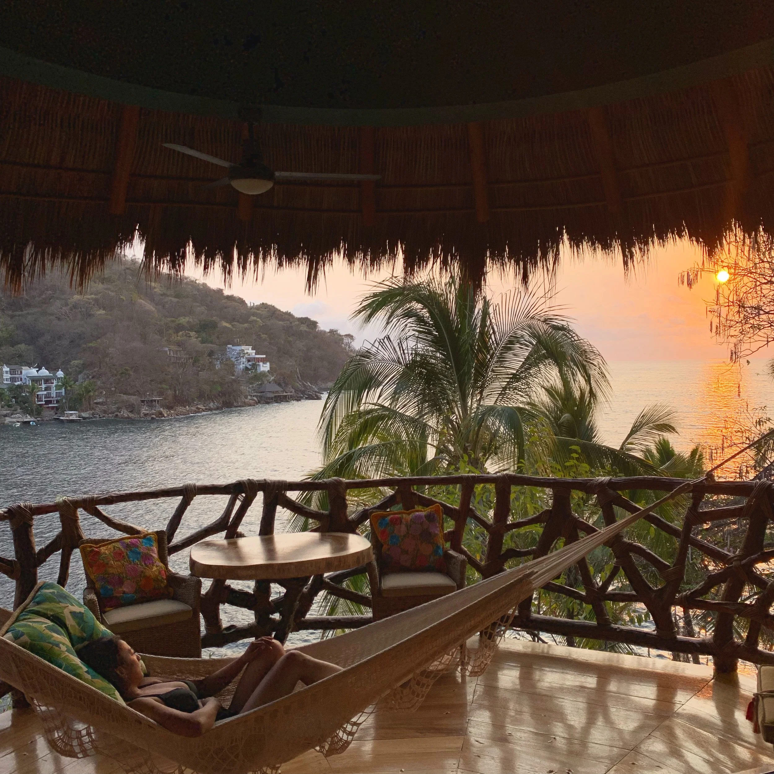 A person relaxing on a hammock with colorful pillows, overlooking a scenic view of water, hillside with homes, and a sunset sky in the background, under a thatched roof structure.