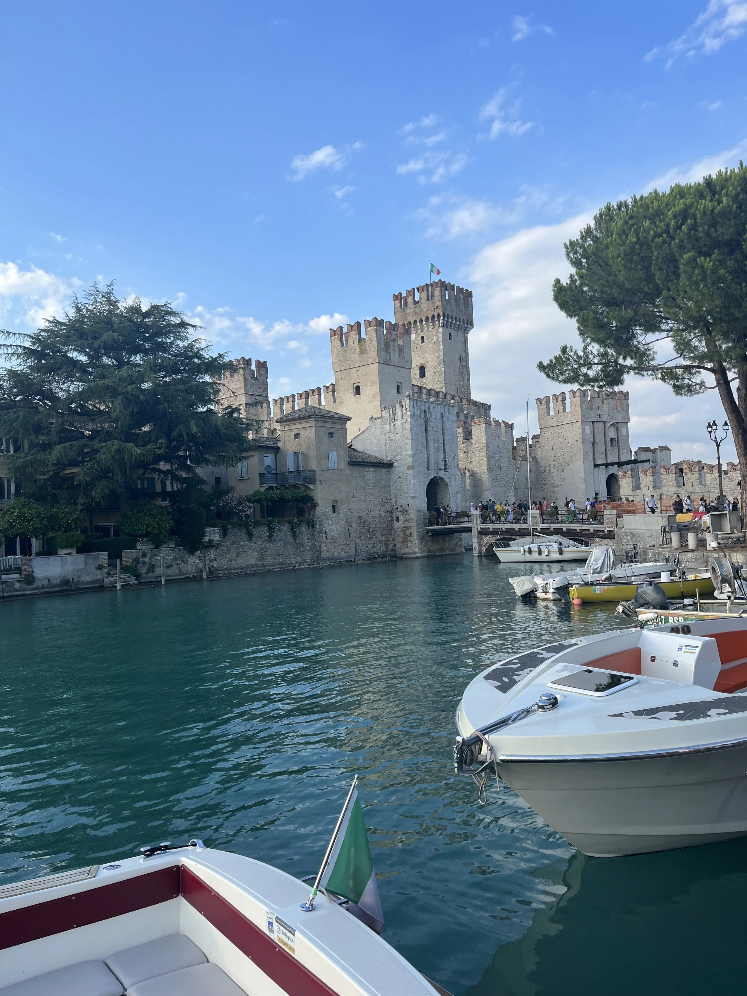 A scenic view of a historic stone castle with multiple towers and battlements, boats docked along the water, lush green trees, and a bright blue sky with some clouds.