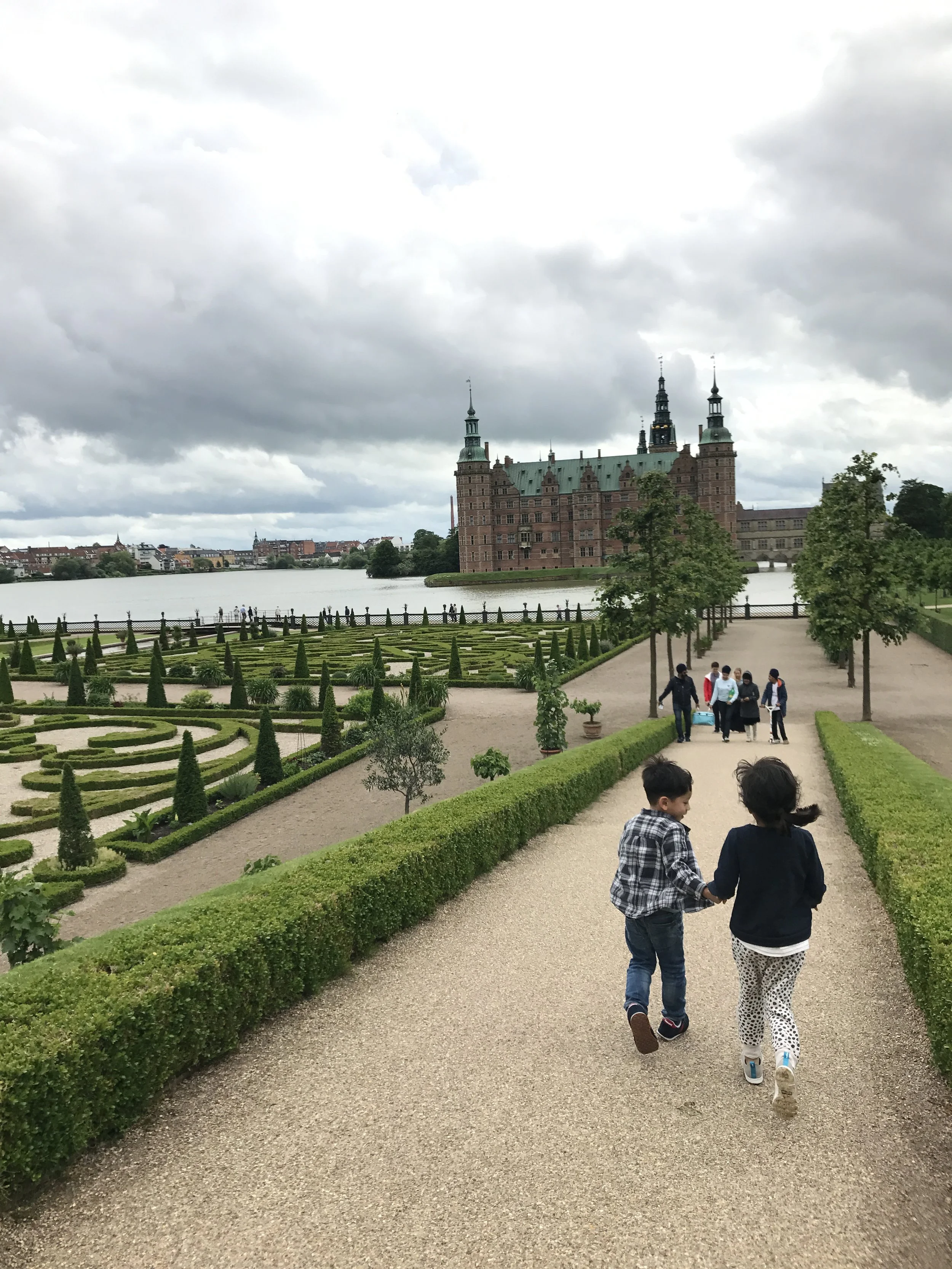 Kids walking on a gravel path with manicured gardens and a historic castle in the background, overcast sky.