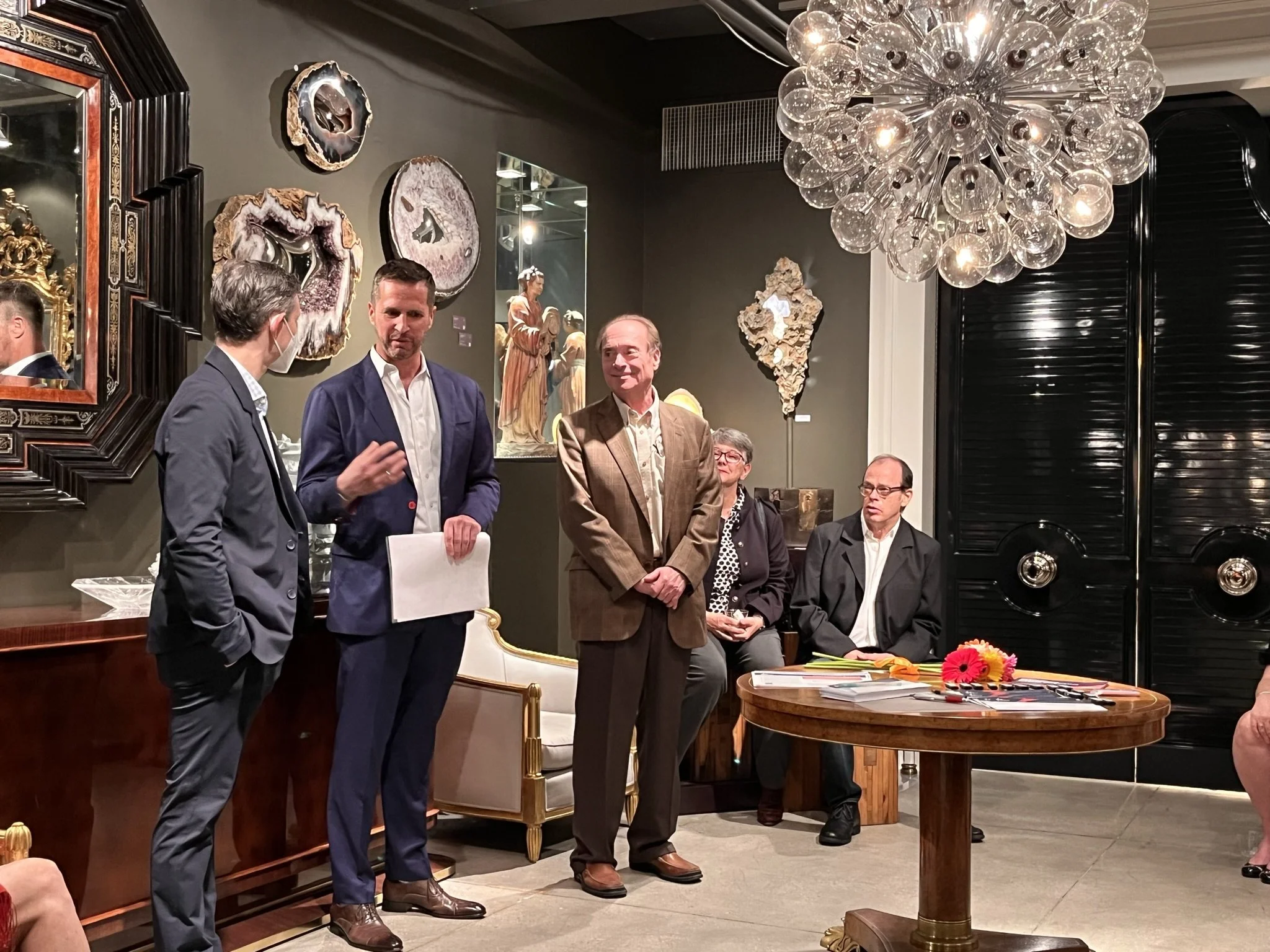 Five men and one woman are gathered in a formal setting with art on the walls and a large chandelier overhead. Some men are engaged in conversation, while others are seated or standing around a table with flowers and papers.