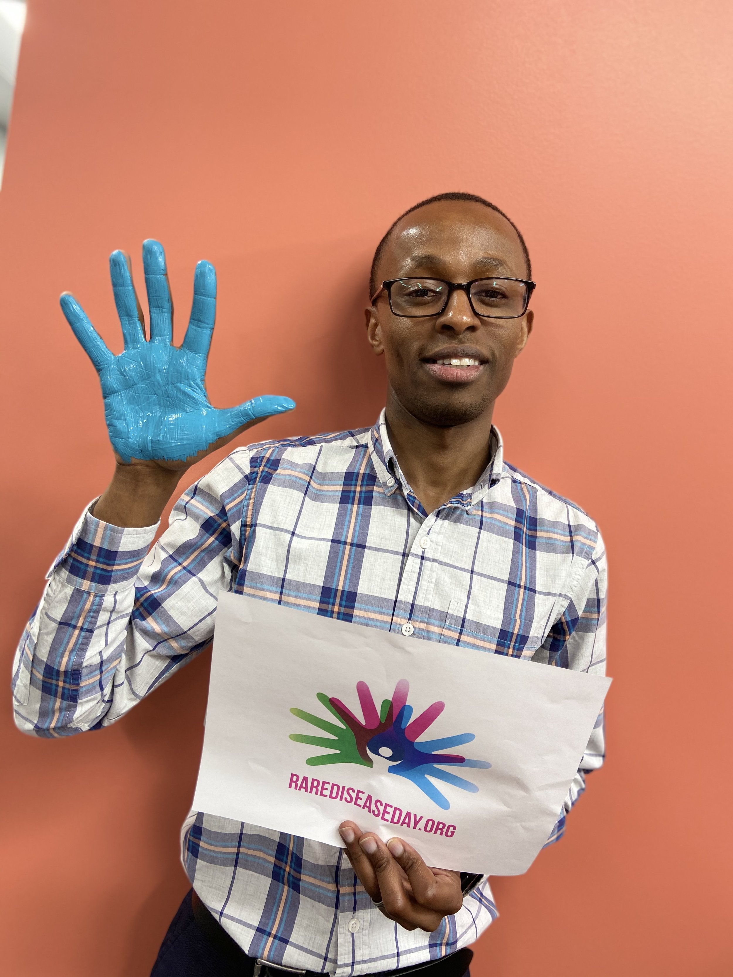 A man with glasses wearing a plaid shirt holds a sign with a colorful logo of multiple hand prints forming a peacock and the text 'RAREDISEASEDAY.ORG'. He is smiling and waving with his other hand, standing against a pink wall.