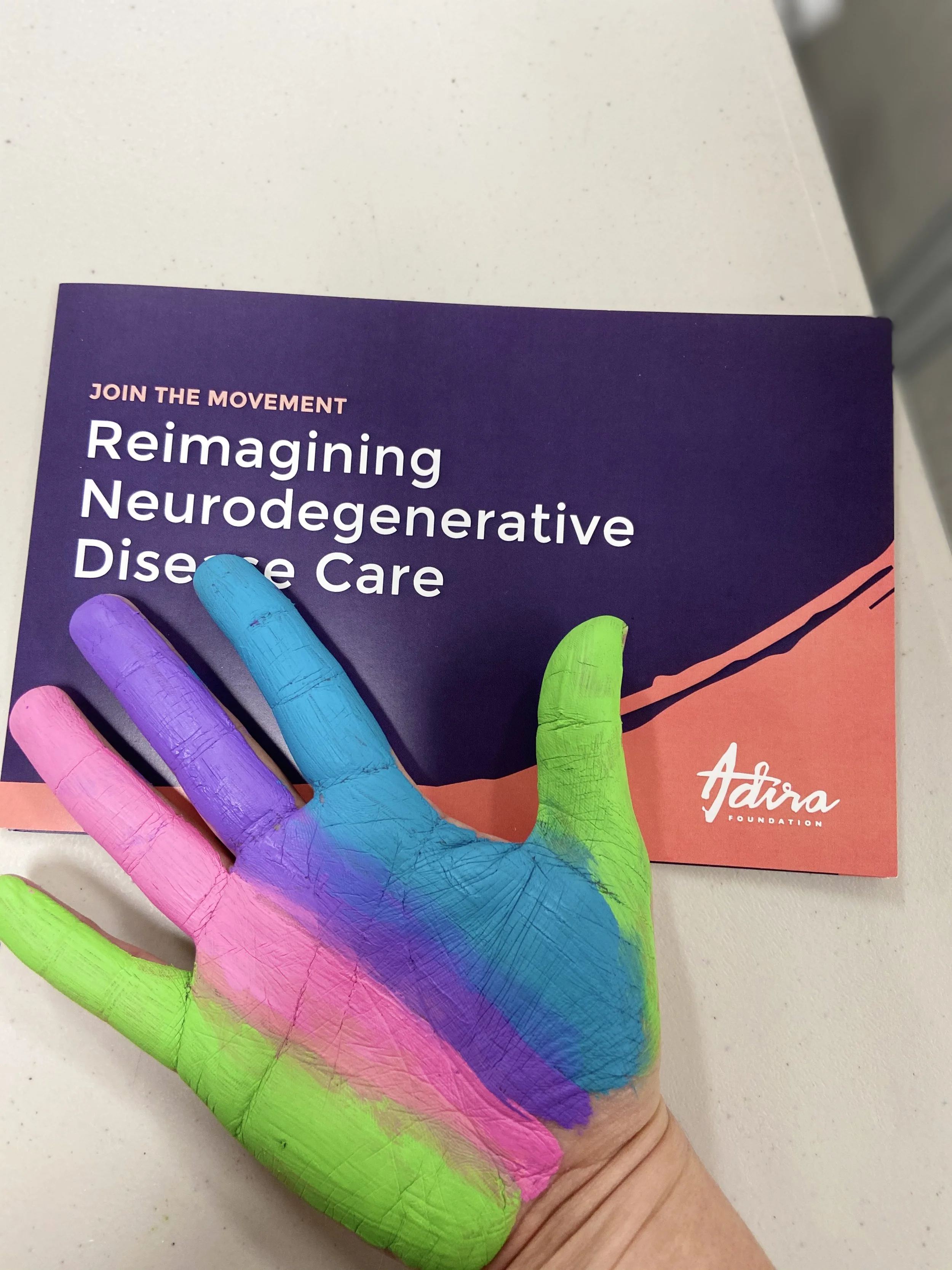 A person's hand painted with rainbow colors, holding a brochure for a neurodegenerative disease care organization called Atara Foundation, on a white surface.