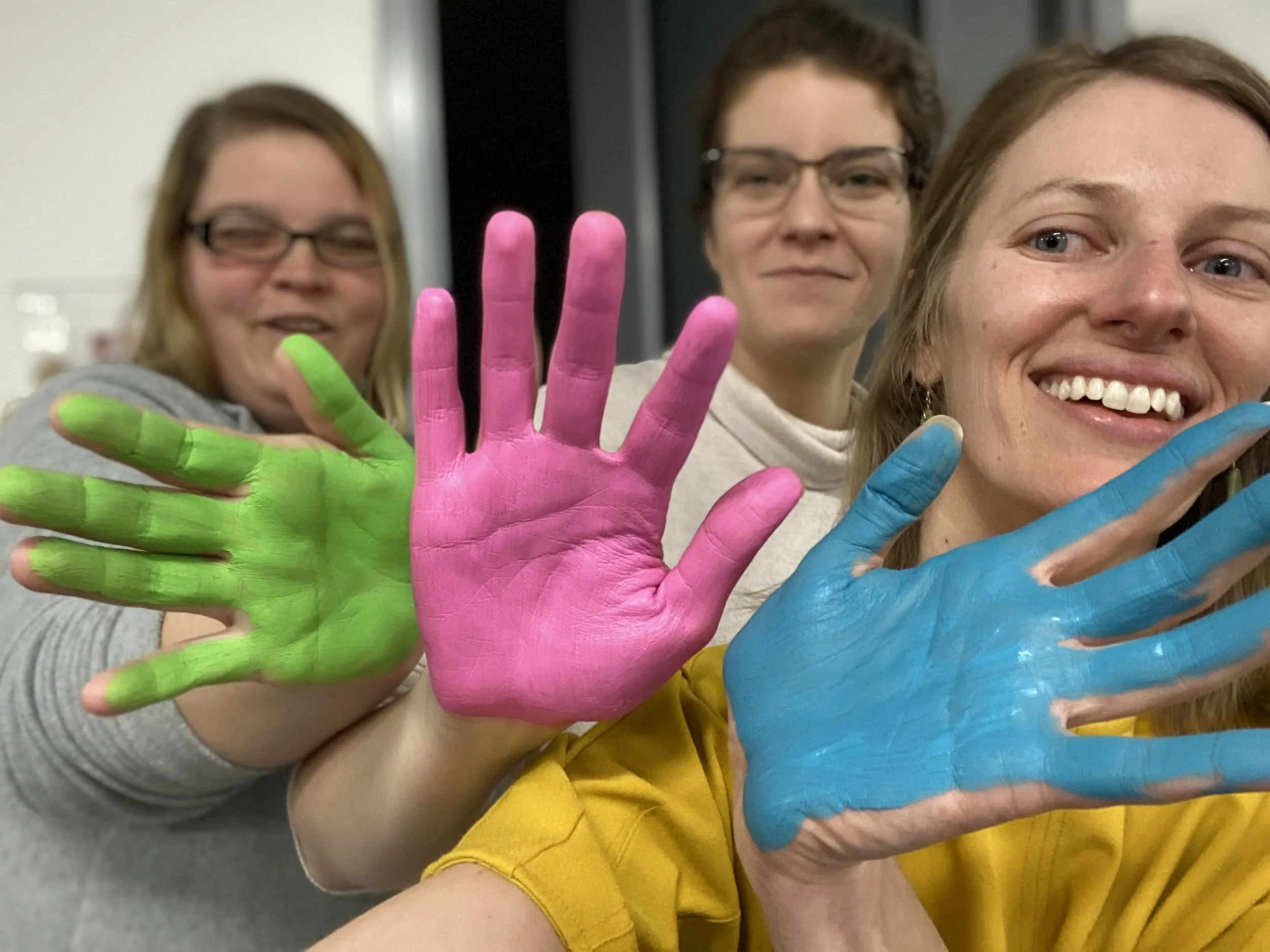 Three smiling women with painted hands showing pink, green, and blue paint. They are close together, smiling at the camera, with a background of an indoor setting.