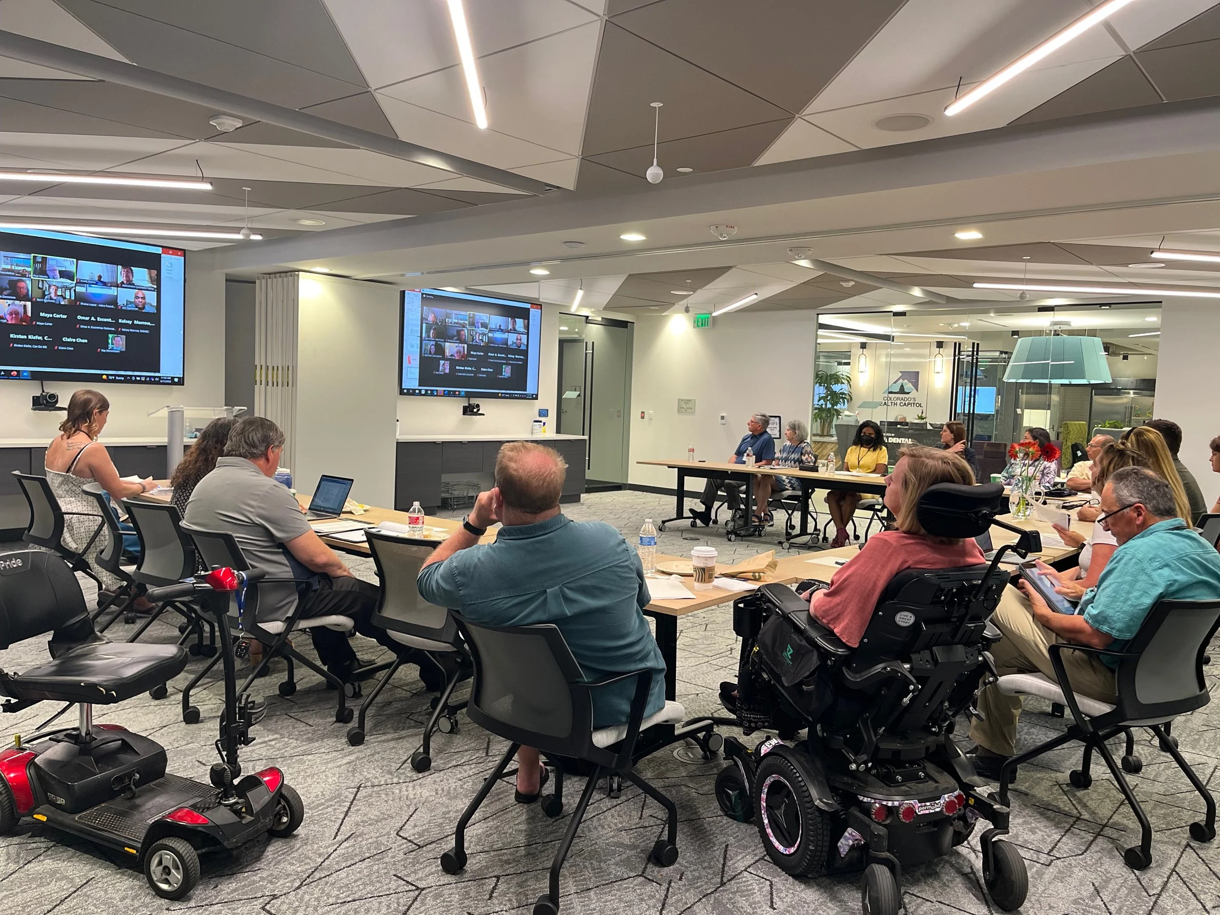 A diverse group of people attending a meeting or conference in a modern, well-lit room with multiple large screens showing virtual participants.