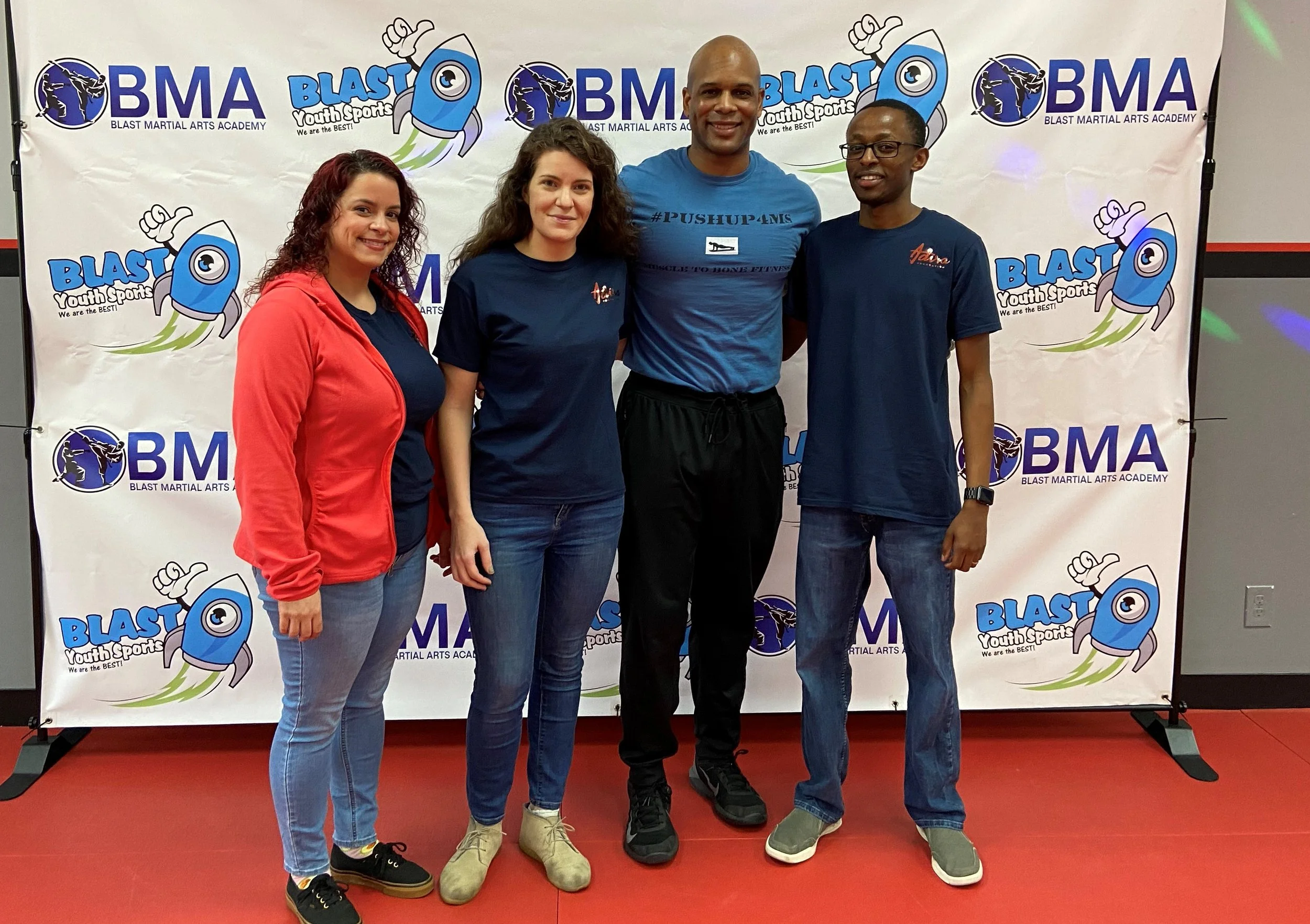 Four people standing in front of a backdrop at a martial arts event. The backdrop displays the logos of BMA Blast Martial Arts Academy and Youth Sports, with illustrations of a rocket. The group includes three women and one man, all smiling and dressed in casual sportswear.