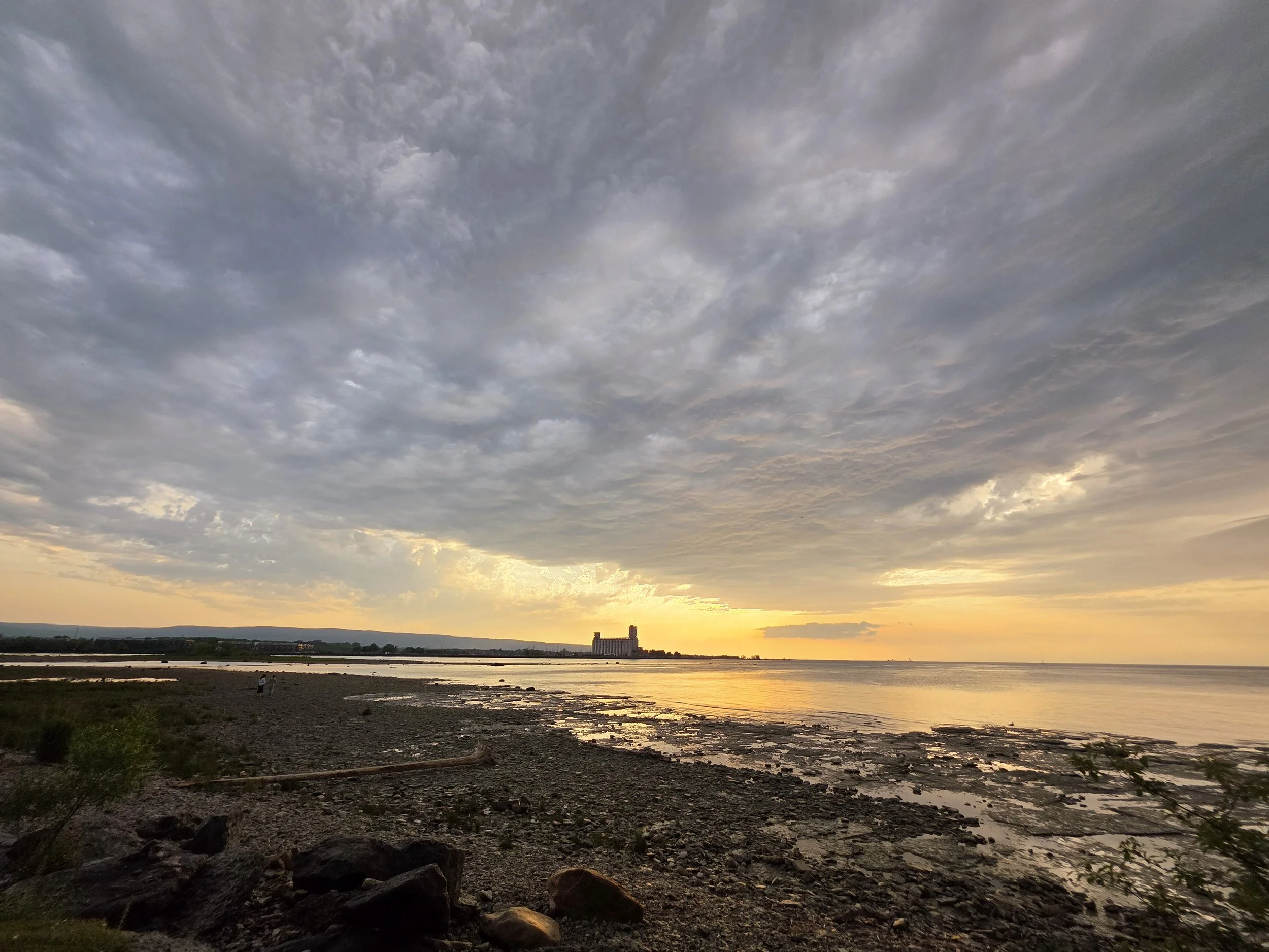 Sunset over a rocky shoreline with a partly cloudy sky and a distant building on the horizon.