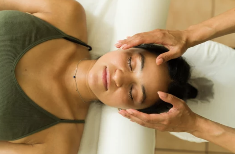 Young woman receiving a head massage while lying on her back with eyes closed in a calming setting.