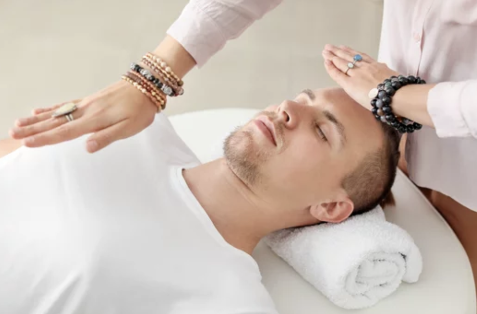 A person receiving a massage or healing treatment, lying on a massage table with their eyes closed, while another person places their hands above the person's head.