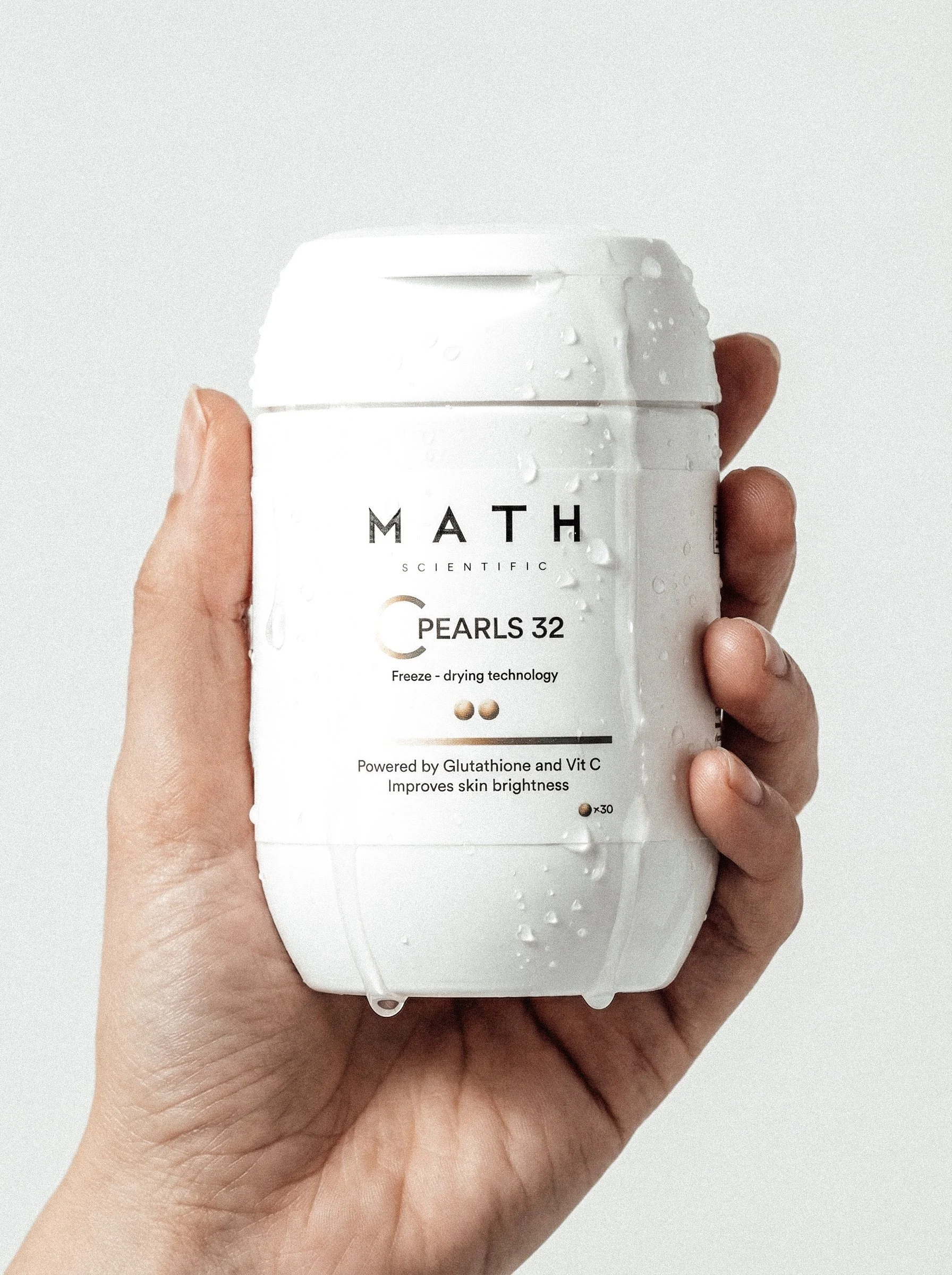Hand holding a white supplement bottle labeled 'Math Scientific Pearls 32' with water droplets on it. The label mentions freeze-drying technology, vitamin C, and skin brightness benefits.