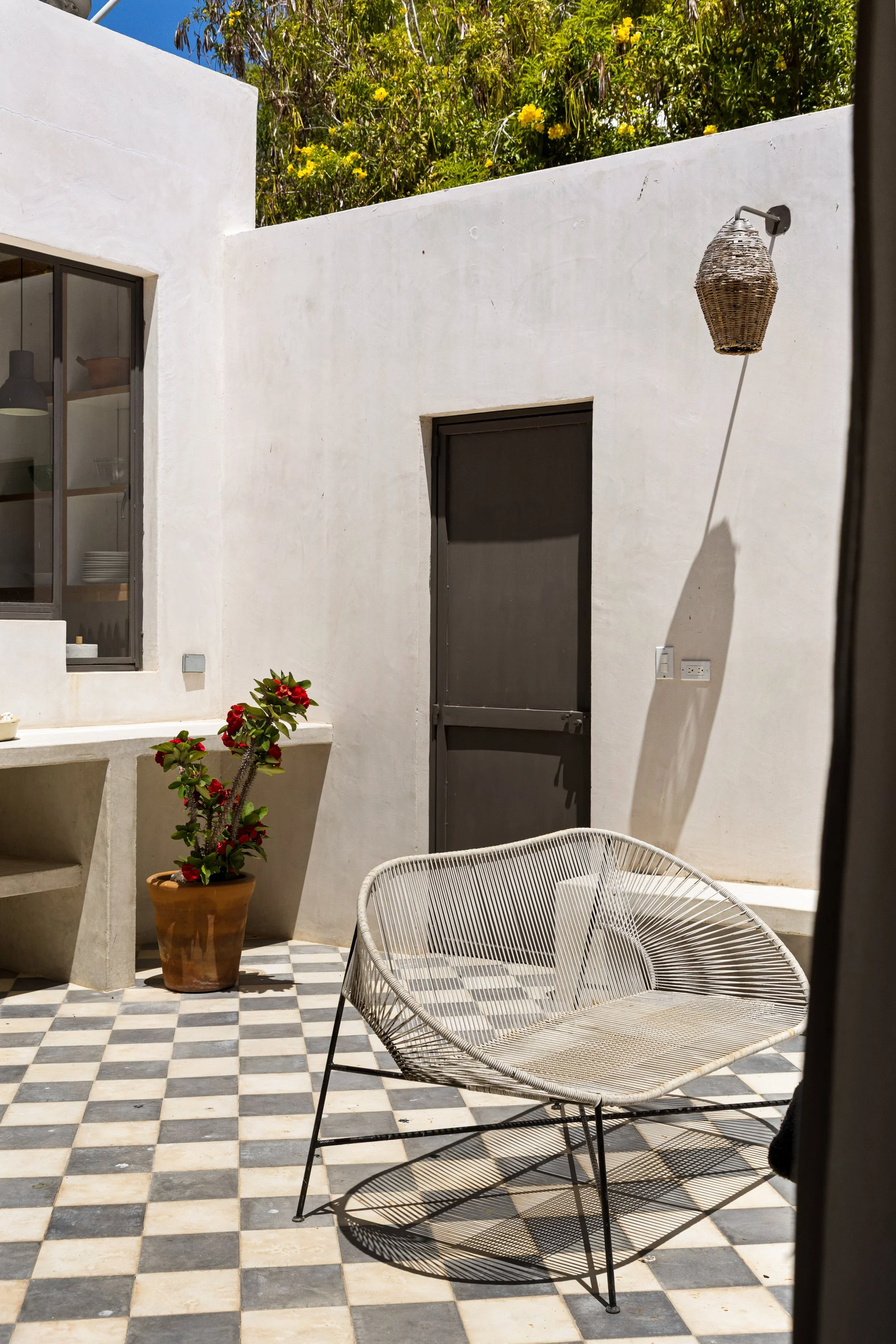 A white outdoor patio with checkered black and beige tiles, a white wire chair, a potted red flowering plant, a white built-in shelf, and a black door, with a white wall and yellow flowers in the background. A wicker outdoor lamp is mounted on the wa