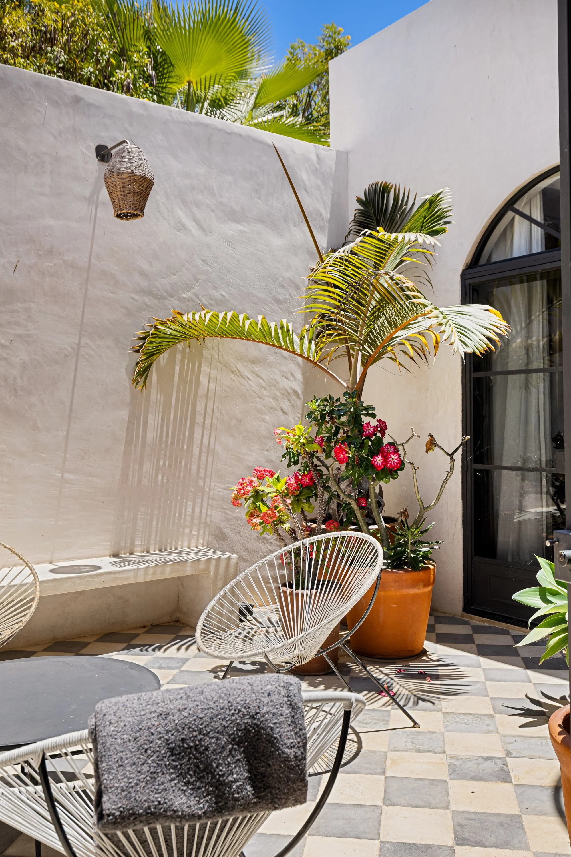 A sunny outdoor patio area with white walls, potted plants, a pair of white modern chairs, and a black round table. The patio has checkered tile flooring and a door with a rounded top window.