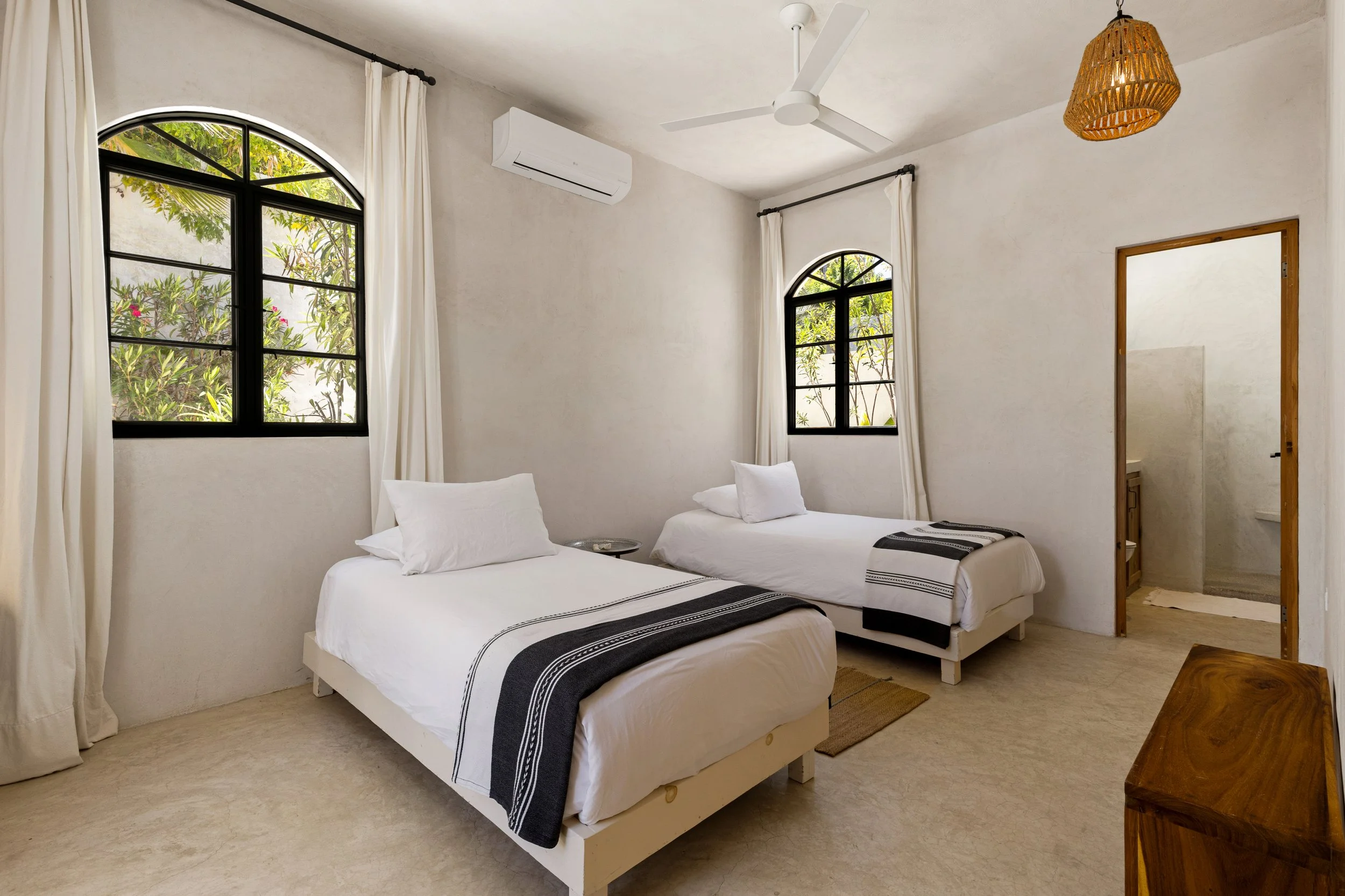 A bedroom with two white beds with black and white striped blankets, white pillows, and white sheets, set against a wall with two large arched windows with black frames and white curtains. There's a ceiling fan, an air conditioning unit, and a hangin
