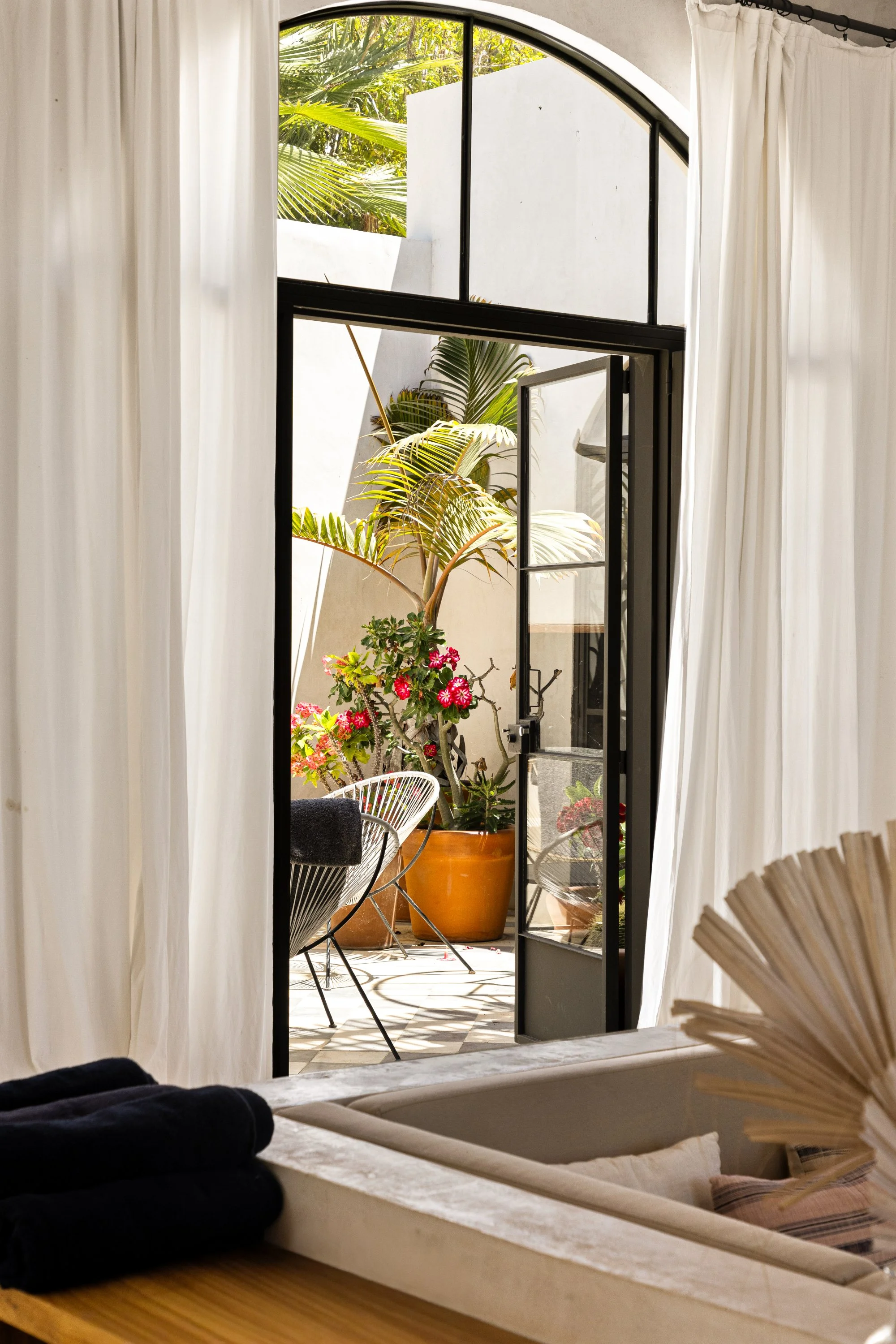 A view from inside a room looking out through an arched door with black metal framing to a patio. The patio has a large potted plant with pink flowers, a small table, and chairs. The scene outside includes lush green trees and plants, with sunlight i