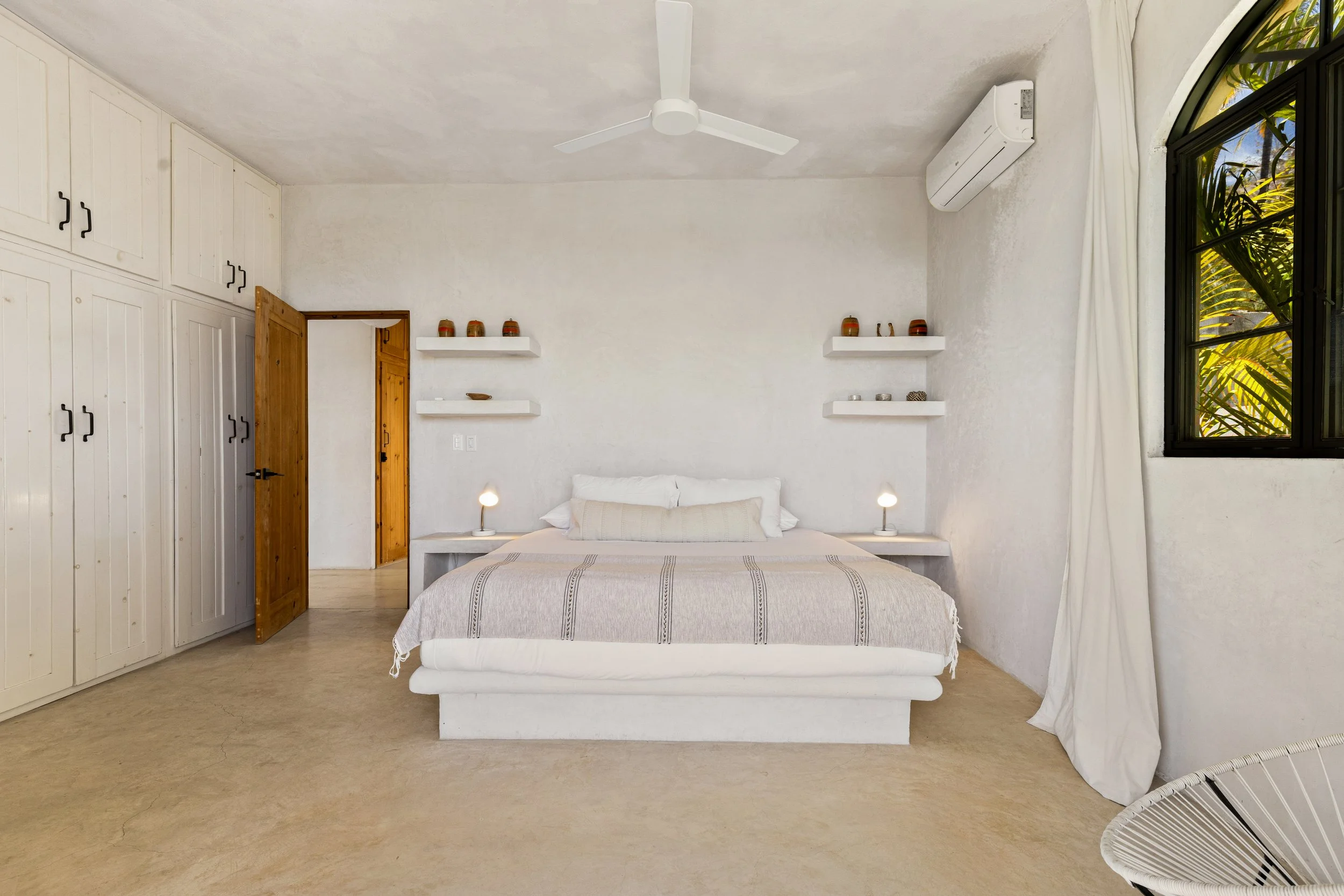 A minimalist bedroom with a white bed, beige blanket, and two small lamps on bedside tables. White ceiling fan, air conditioning unit, white built-in wardrobe, wooden door, and window with greenery outside. Wall-mounted shelves with decorative vases.