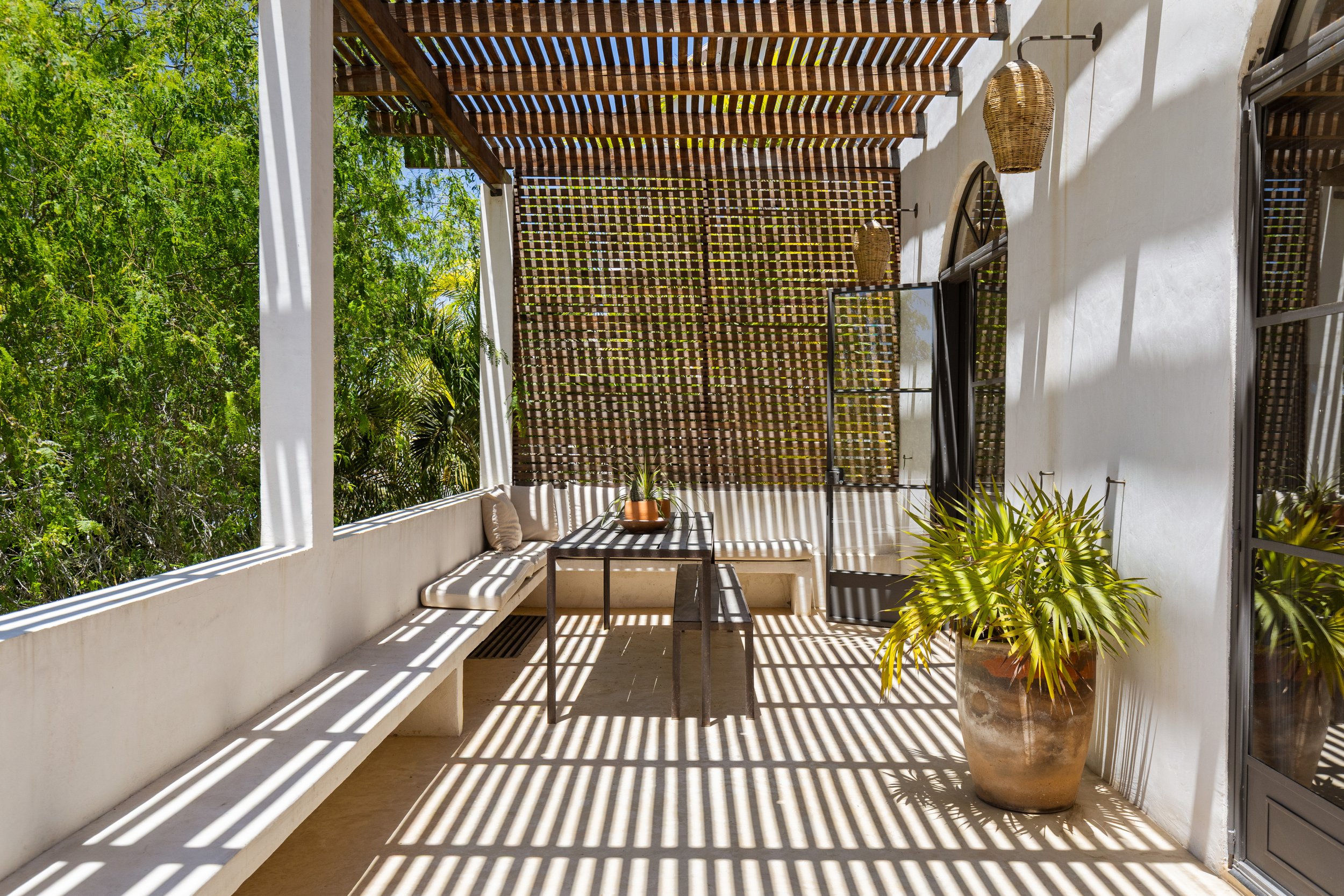 A balcony with a white wall and wooden slats casting striped shadows, green trees in the background, outdoor seating with cushions, a small table with a potted plant, and potted plants near the door.