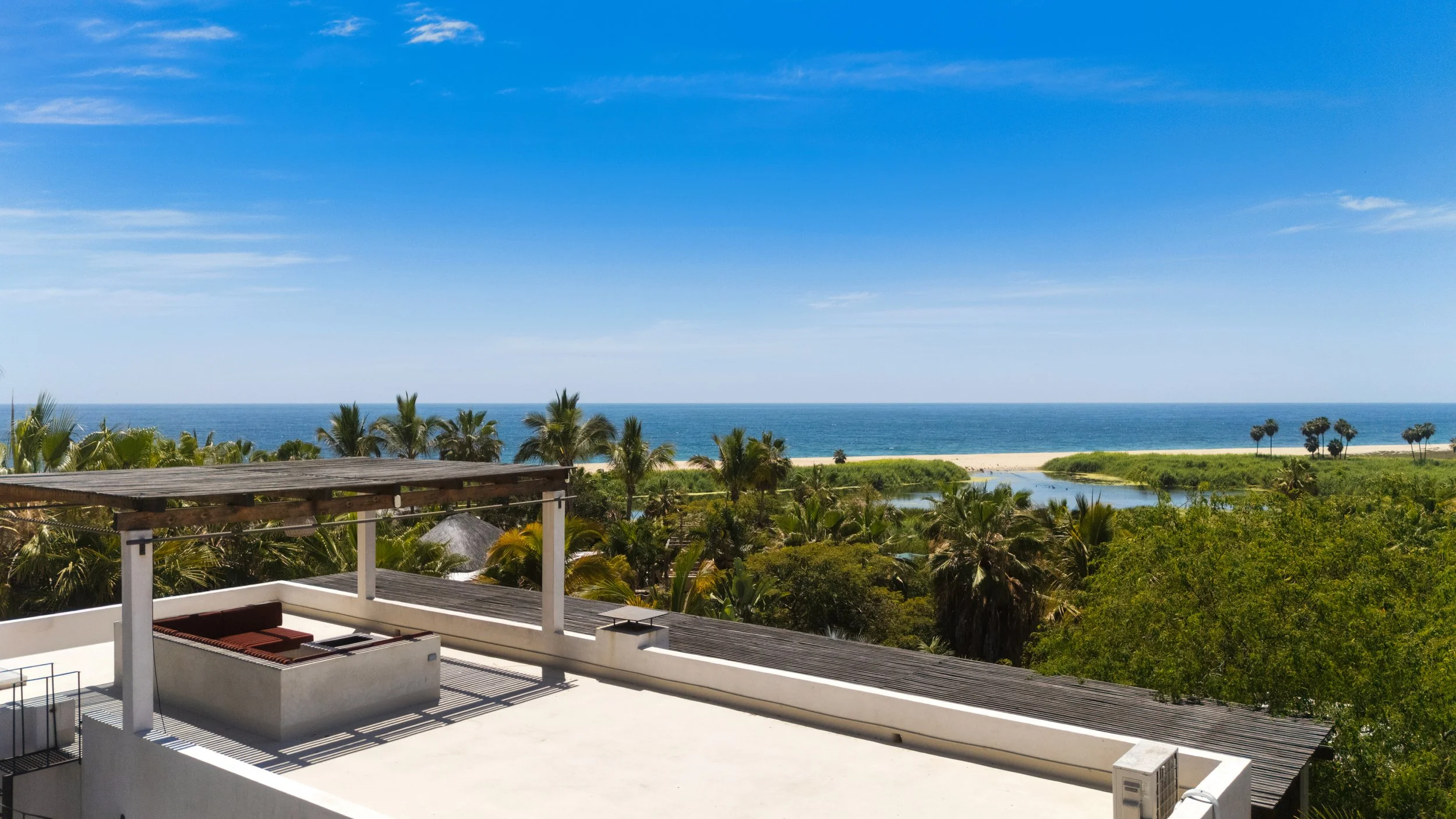 View from a balcony overlooking tropical palm trees, a river, and the ocean under a blue sky.